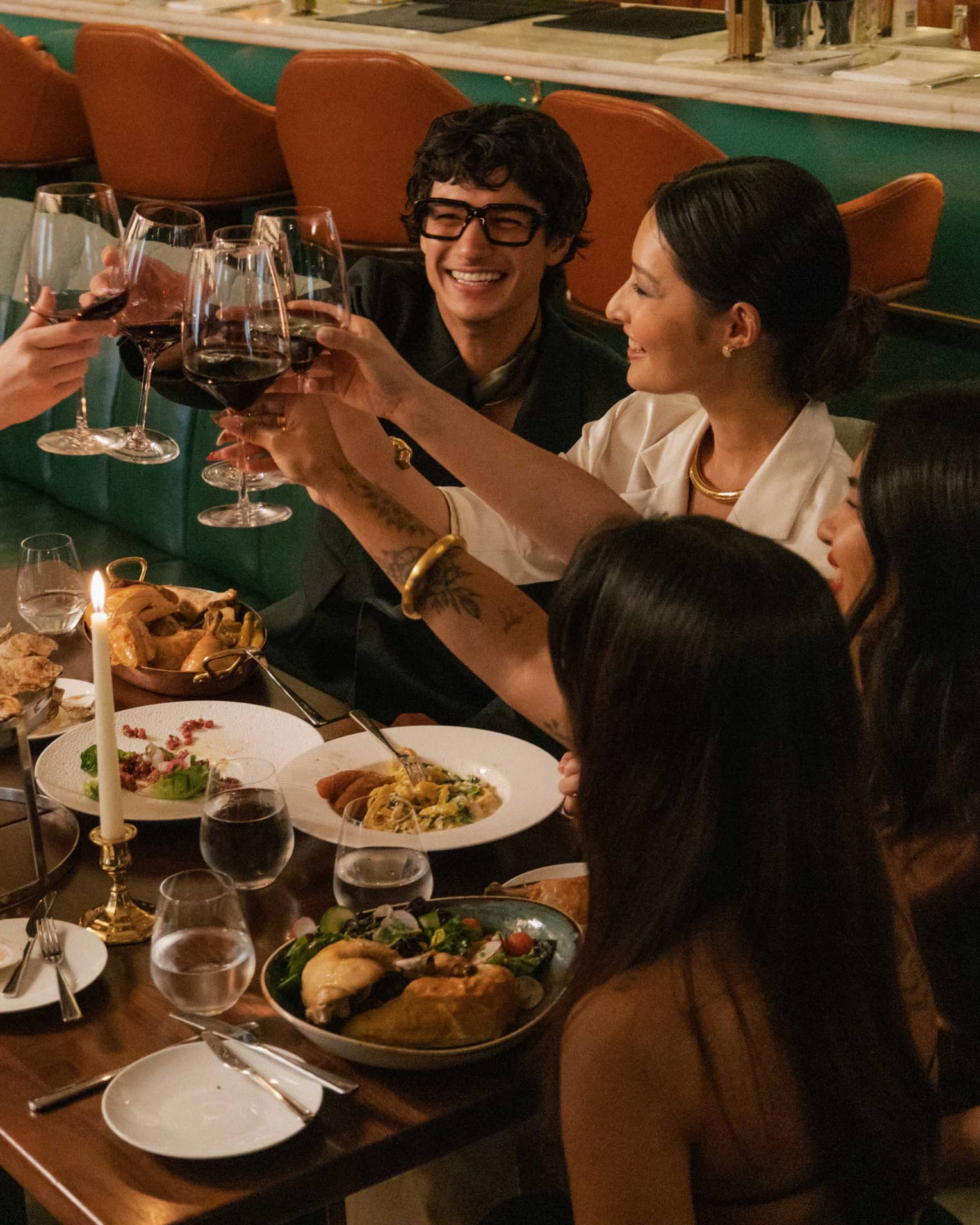 Diners seated in a banquette smile as they clink their red-wine glasses in a toast to a sumptuous candlelit feast.
