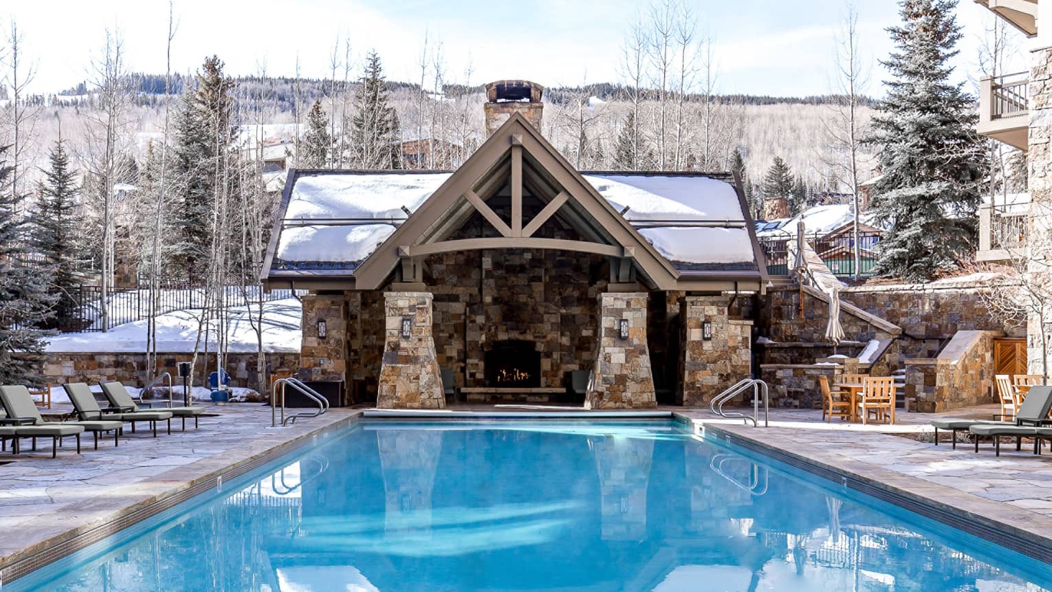 Outdoor pool with lounge chairs, a cabin with a fireplace and pine trees in the background.