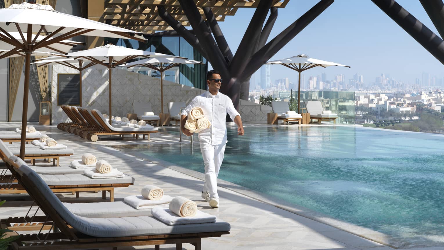 Hotel staff walks past white lounge chairs on outdoor swimming pool deck, carries rolled towels 