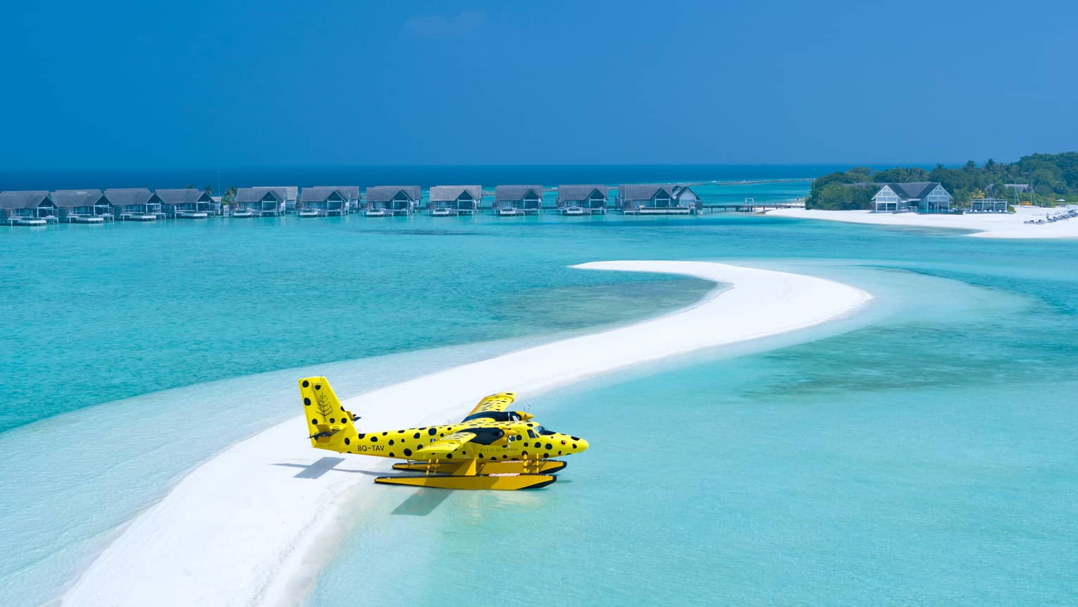 Yellow and black-spotted single-engine plane flies over clear waters of the Maldives, overwater bungalows in distance