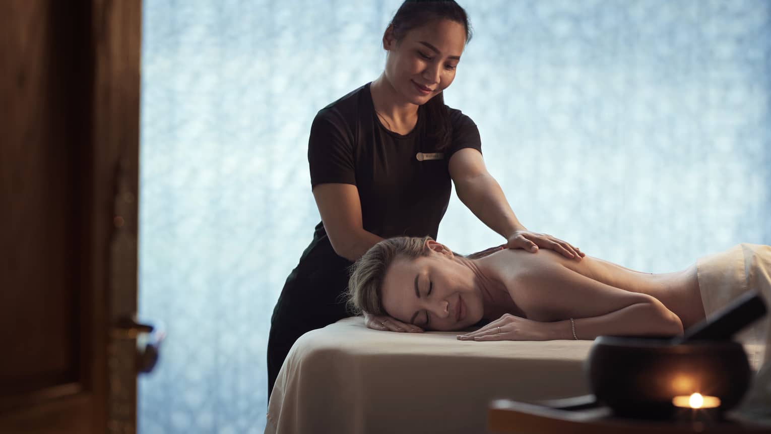 Spa staff massages woman's shoulders as she lays on bed in dark room