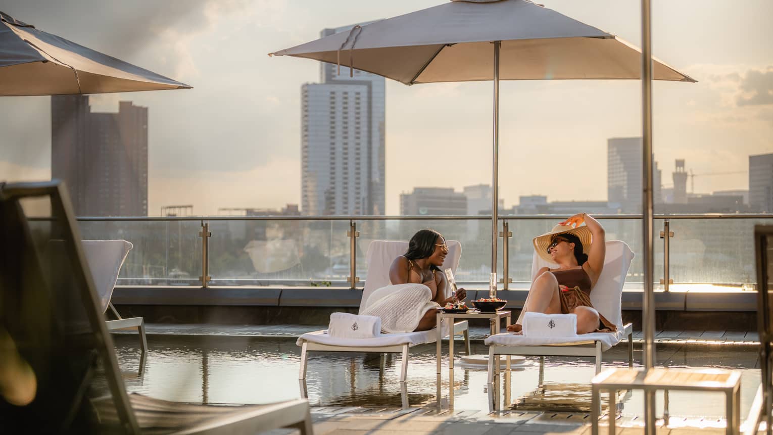 Two guests visit with one another while seated on lounge chairs under a sun umbrella and over a rooftop pool, with city skyline views in the background.