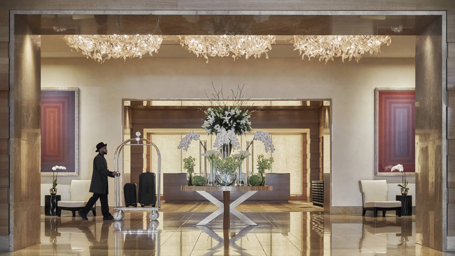 A man in black clothes pushing a trolly of suitcases across a hotel lobby that is lit by gold chandaliers.