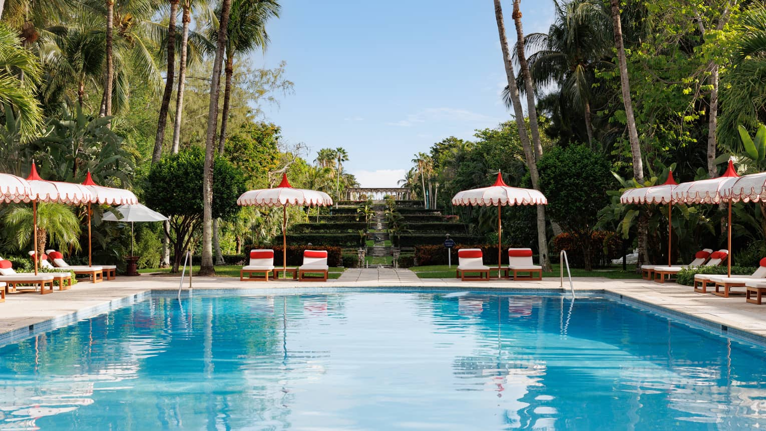 An outdoor pool at a tropical resort, flanked by white and red lounge chairs and umbrellas
