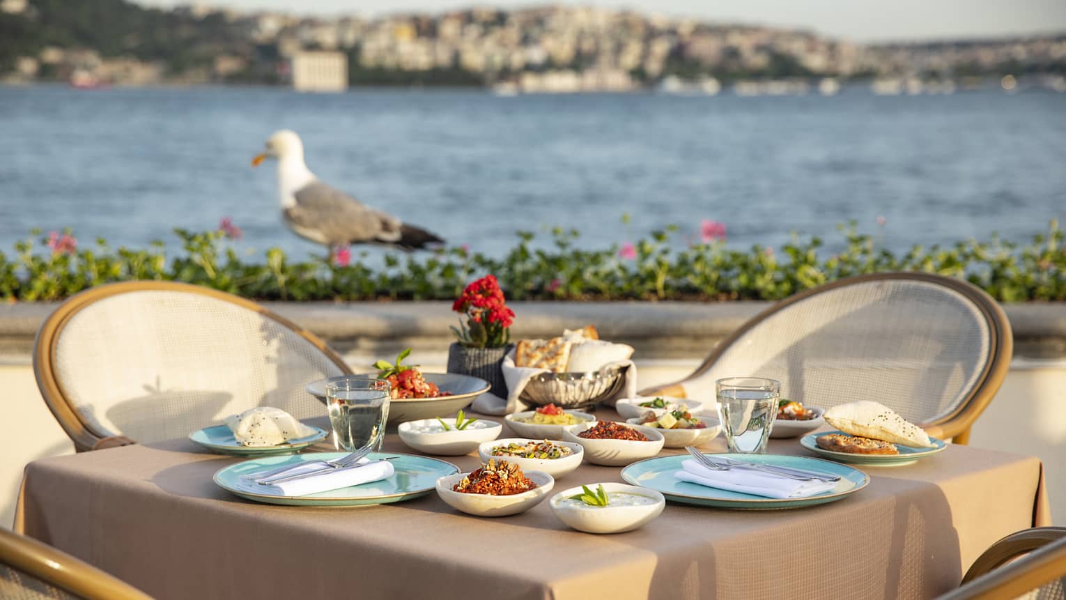 A table set for four with rattan chairs and beige linens, a gull and the Sea of Marmara in the background