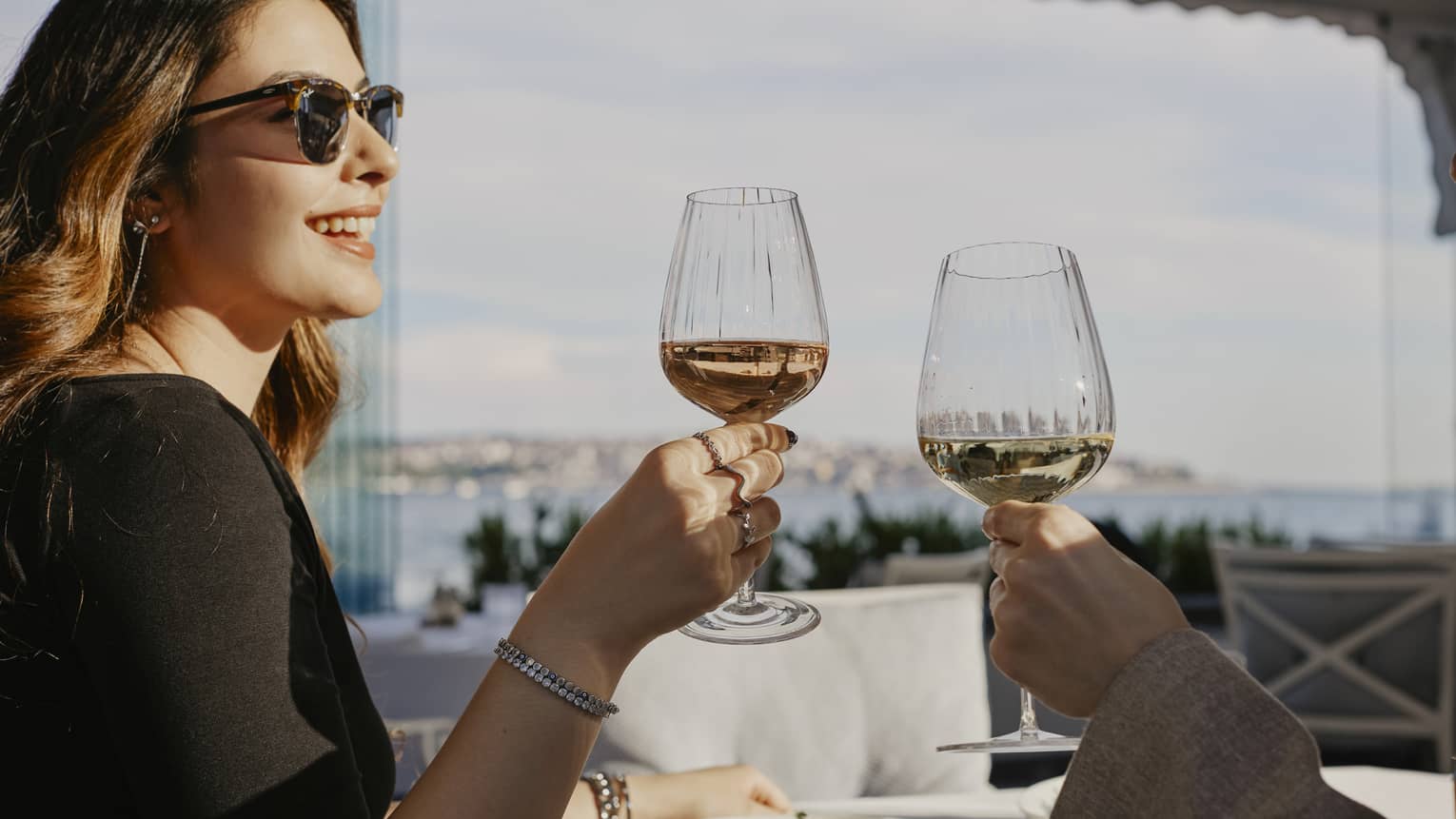 Person wearing sunglasses, smiling while toasting with a glass of wine with another person