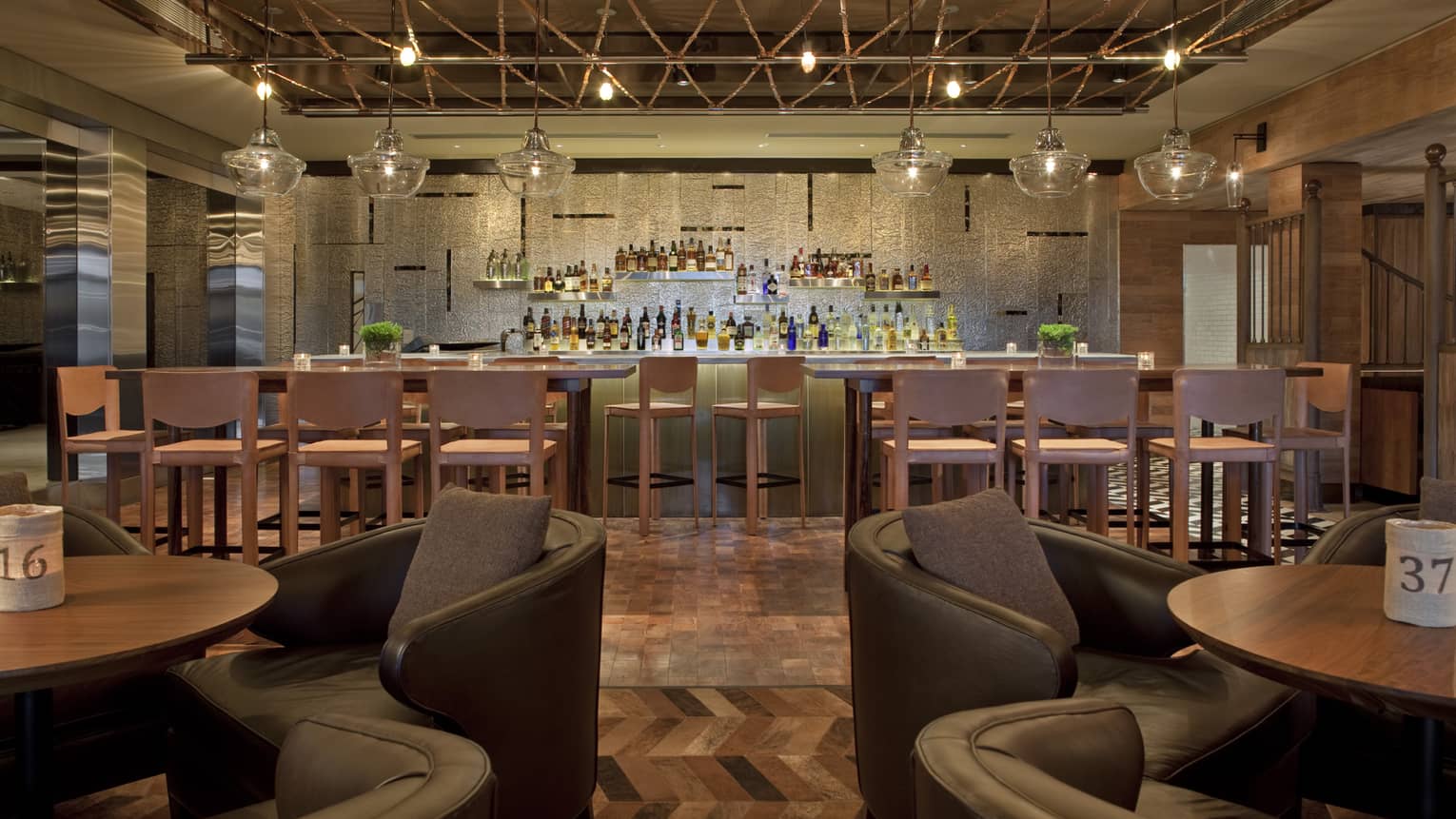 A brown bar with herringbone flooring, glass light fixtures, an exposed wood ceiling, and a spacious bar area with ascending bottles of liquor.