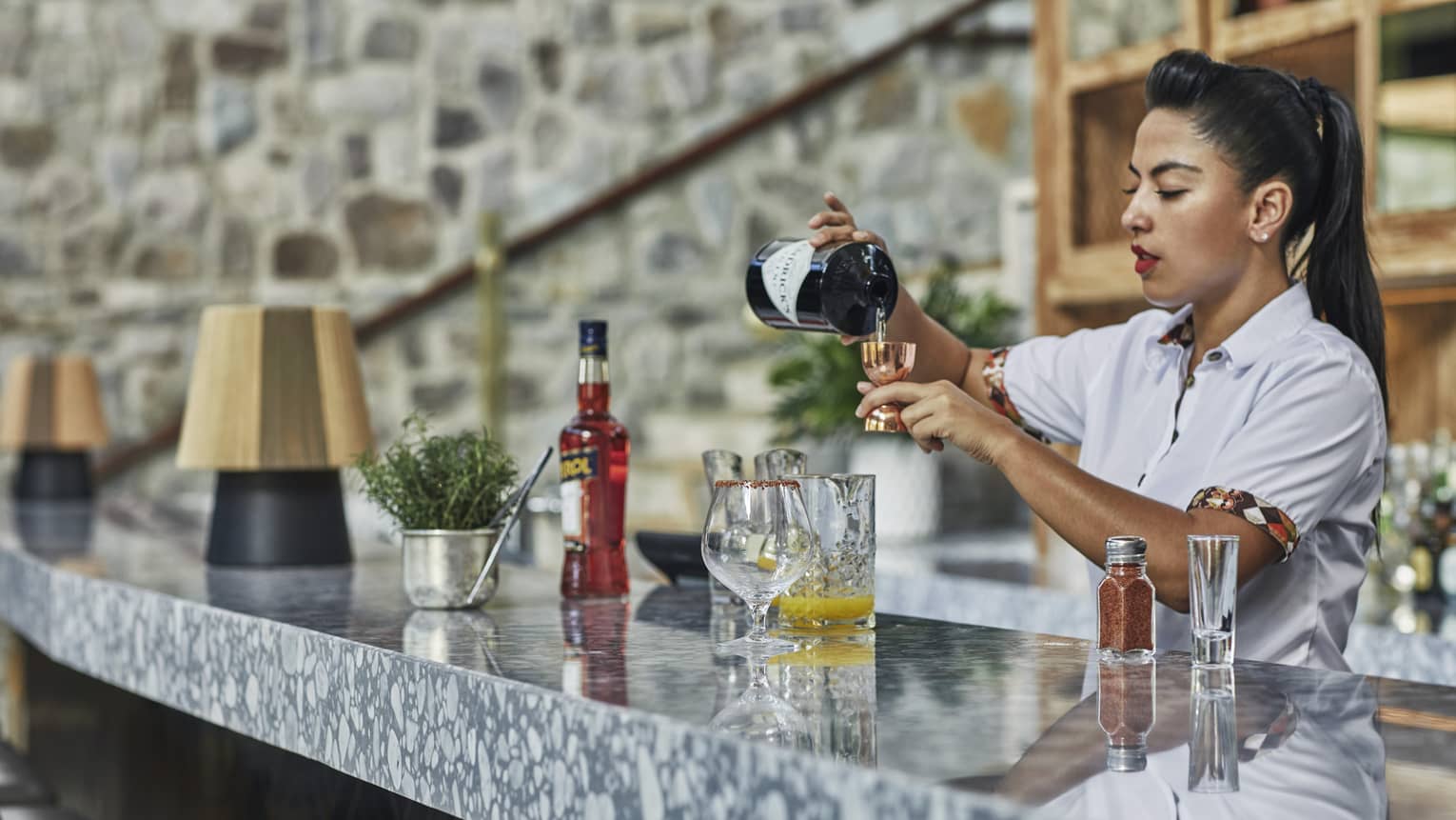 A woman with a high ponytail prepares a drink behind a marble countertop.