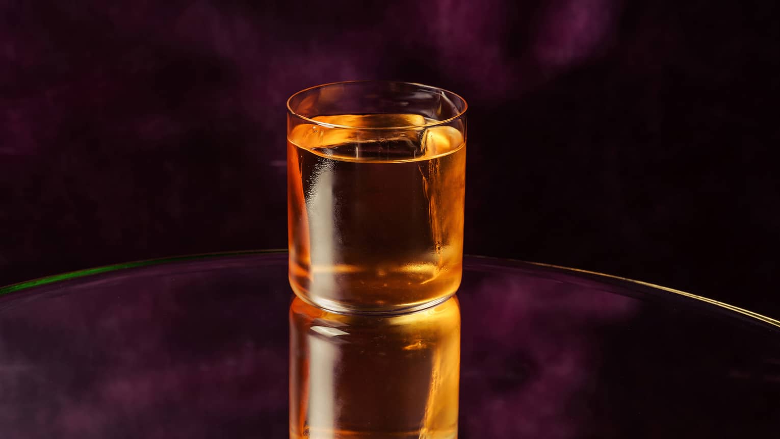 A golden-colored beverage in a rocks glass shown on a dark violet backdrop