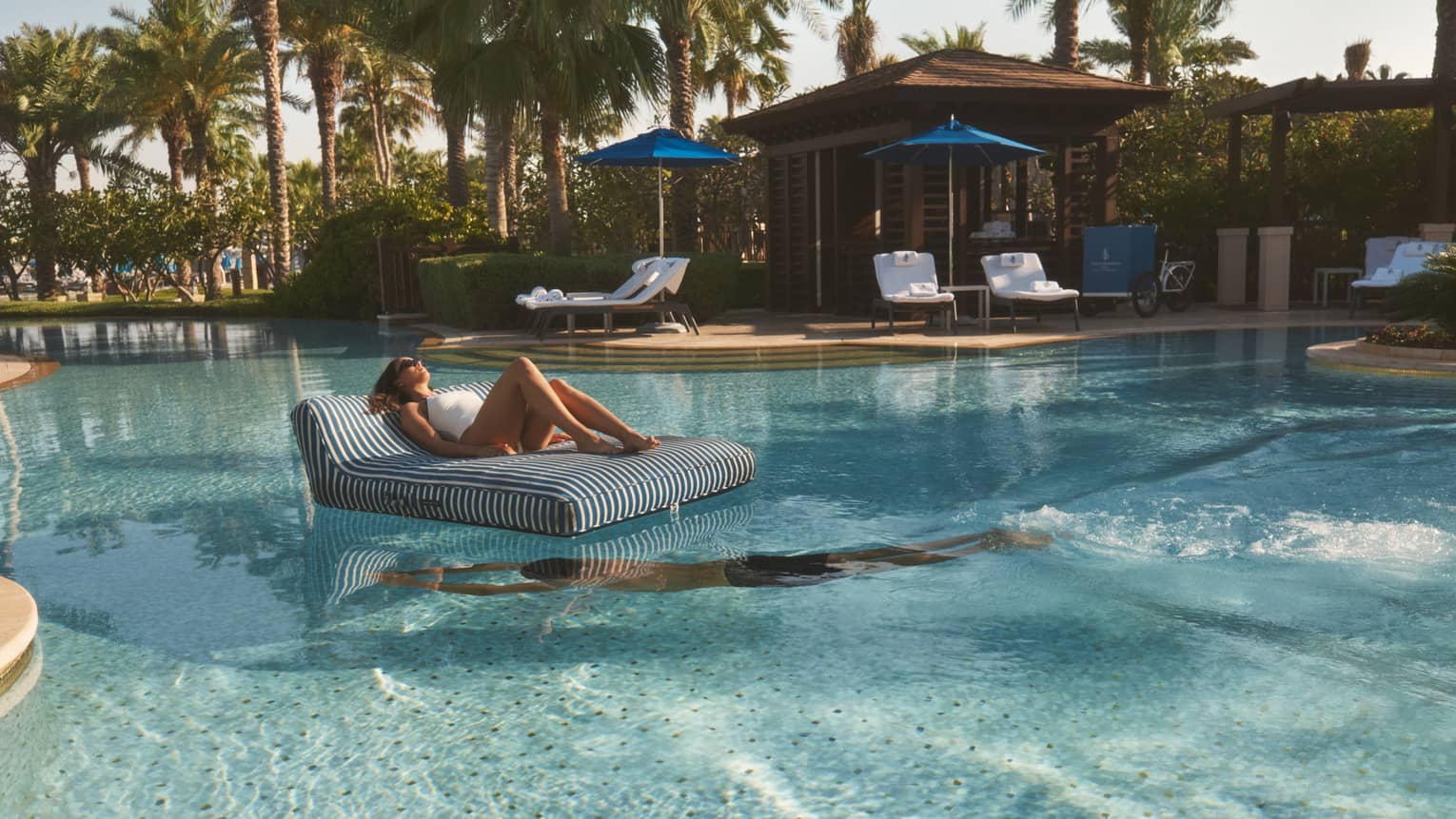 Person lounges on a black-and-white striped floating chair in the middle of a pool as another person swim beneath