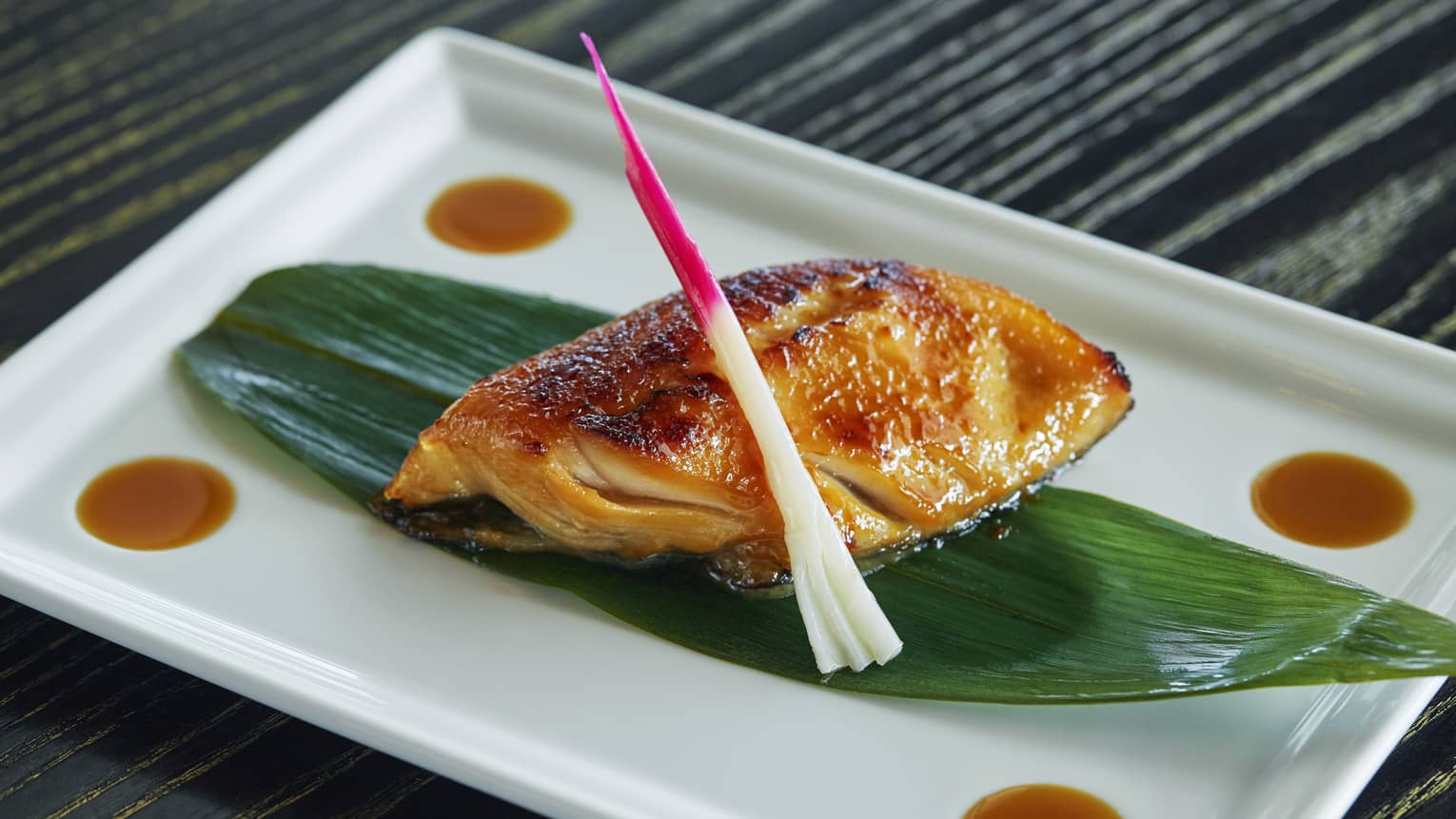 Black Cod Yuzu Miso fish fillet marinated, oven-baked on grilled seafood filet on leaf on white platter