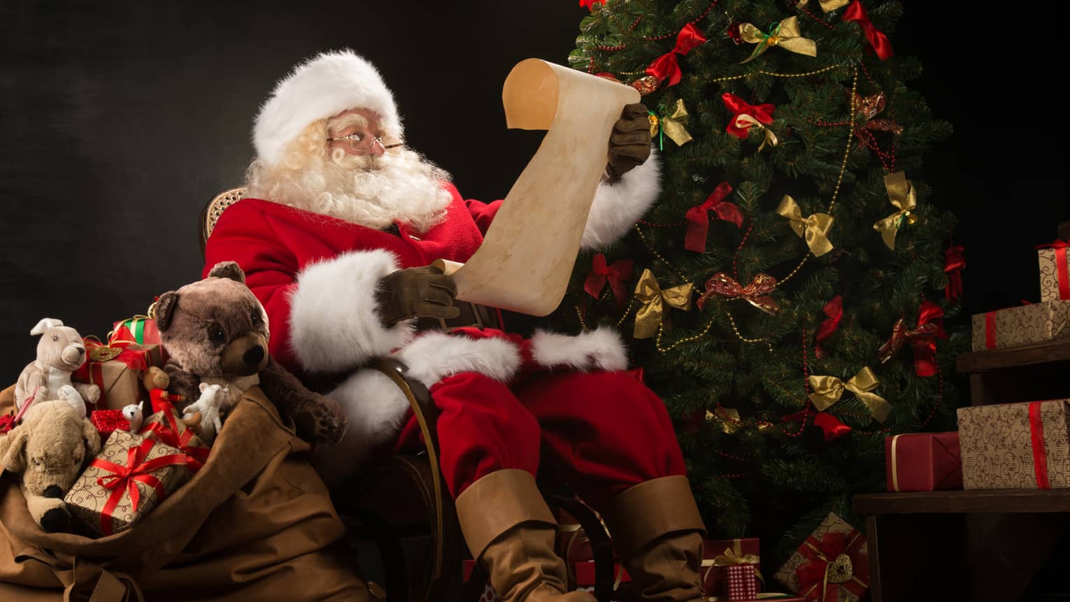 Santa Claus seated in large rocker holding a scroll, flanked by decorated Christmas tree with gifts and overflowing gift bag
