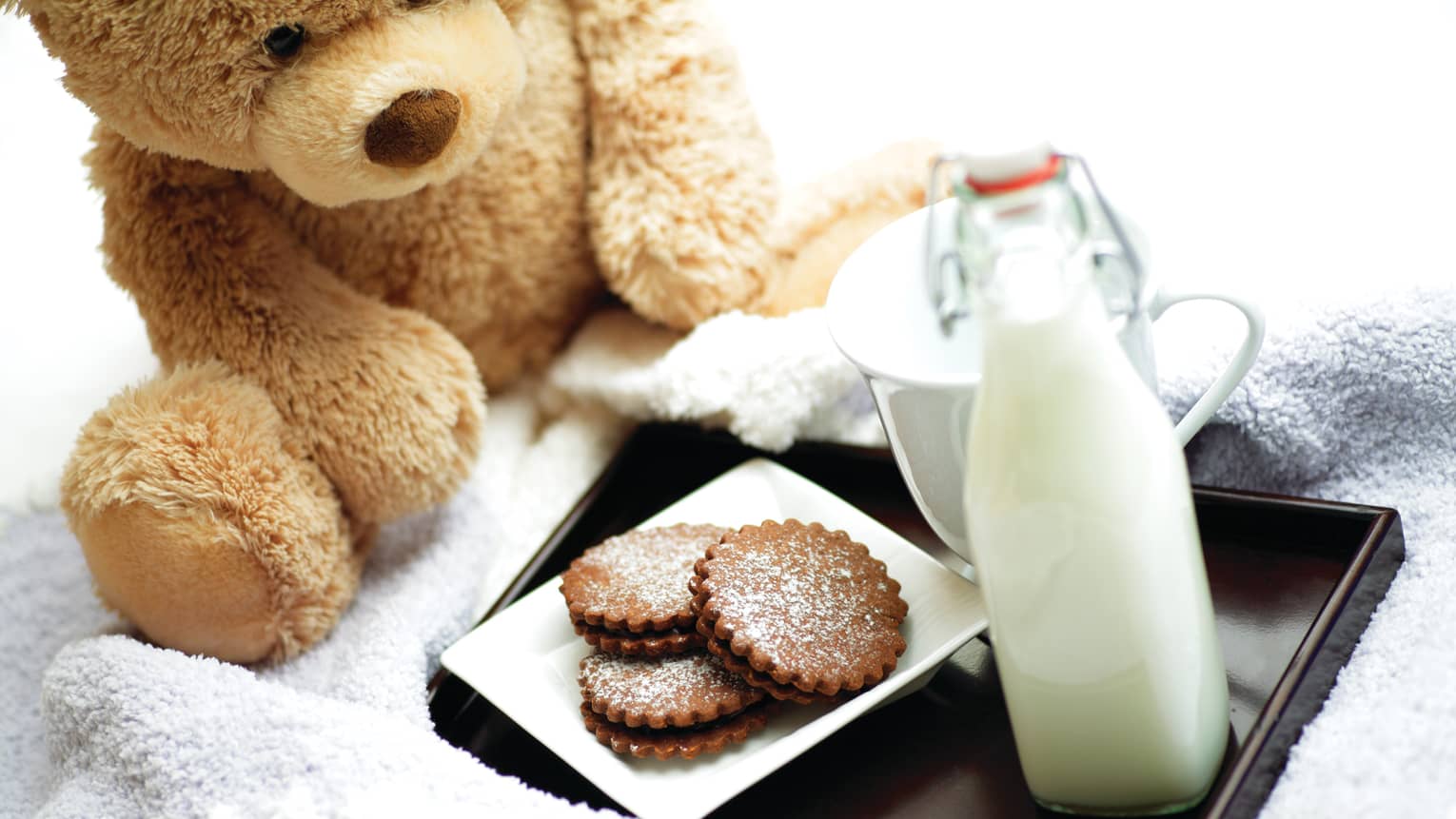 A teddy bear next to a tray of milk and cookies.