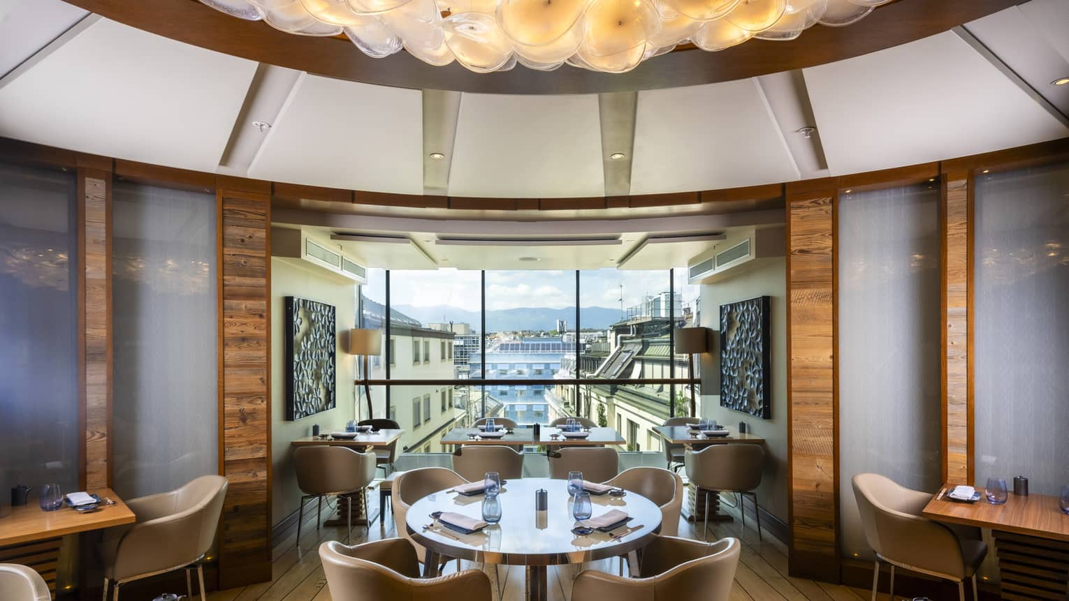 A dining space bright with natural light and light from an artistic ceiling fixture. Large windows are in the background, and the space features earth-toned decor and several small dining tables with upholstered chairs.