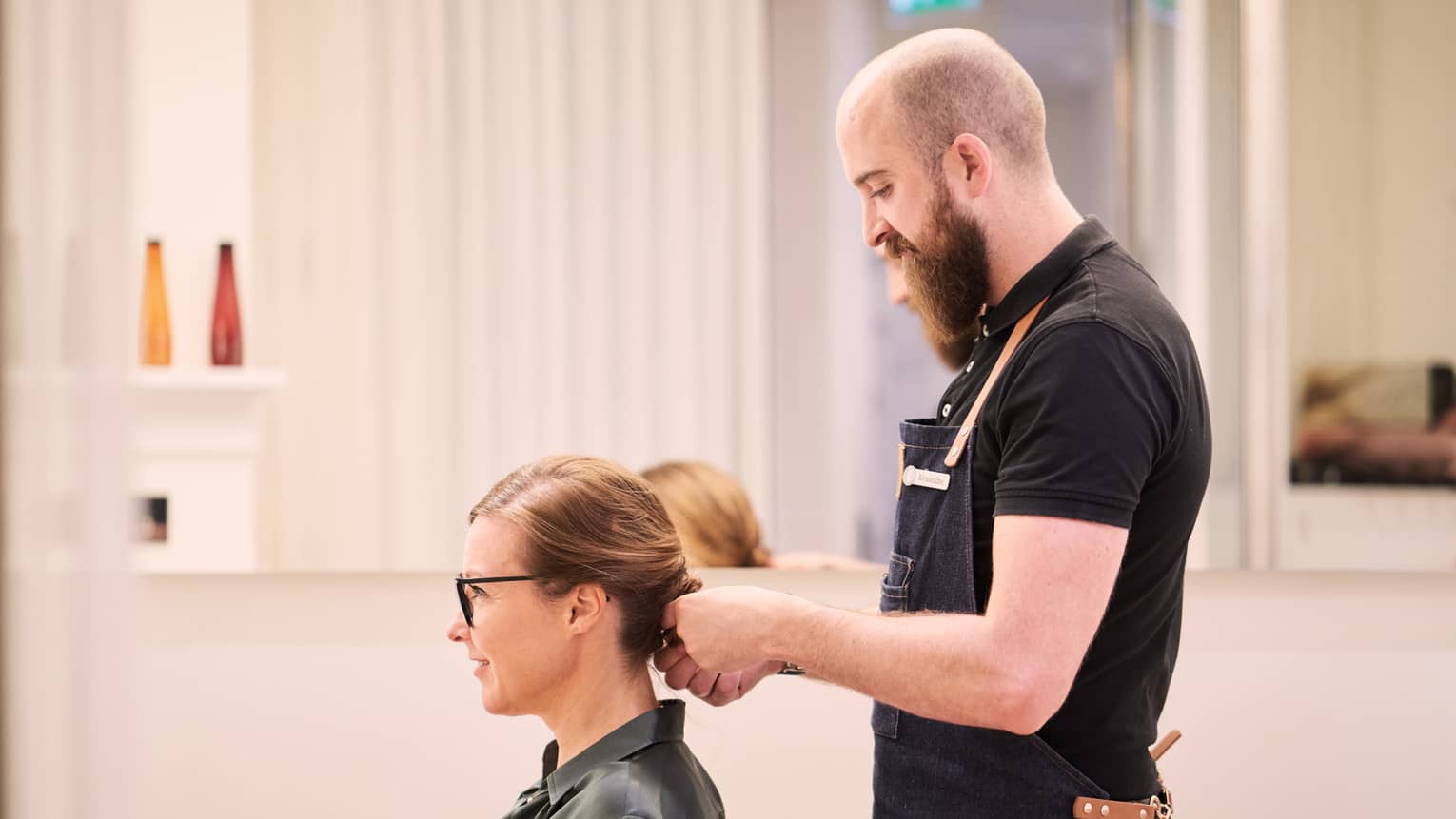 Woman sits in salon chair as hair stylist with long beard holds her hair