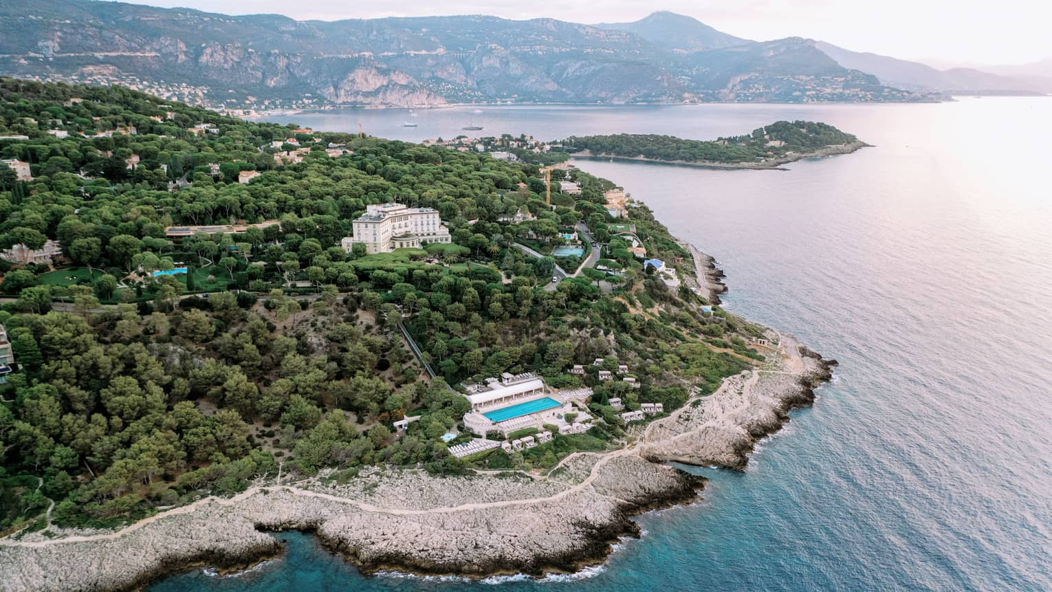 Aerial view of buildings and pools nestled among verdant trees on a sloping hillside along an extensive, rugged coastline.
