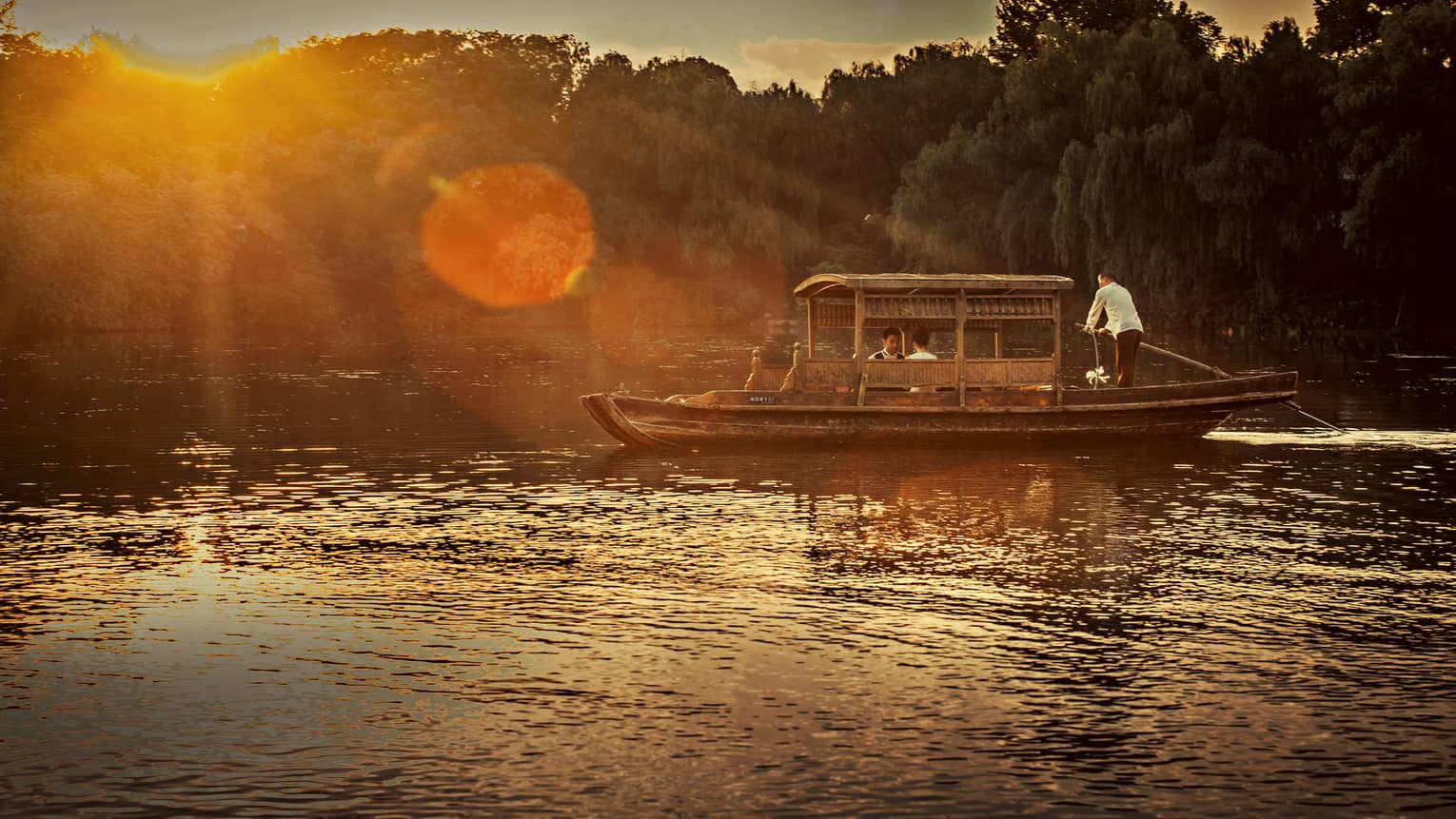 Man steers Chinese wooden rowboat on West Lake, into light of orange sunset
