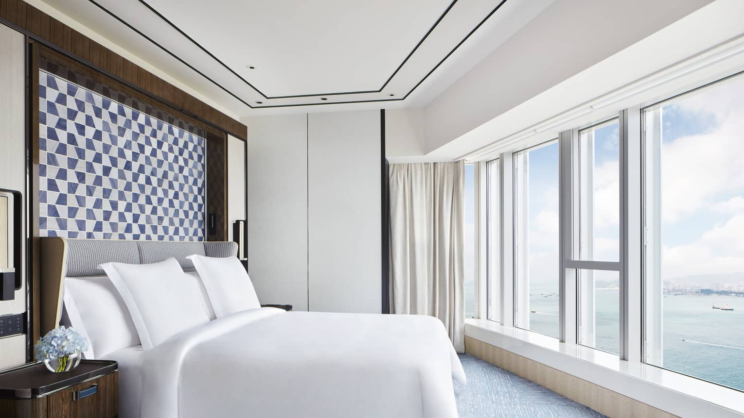 Bright hotel bedroom with large bed, blue geometric headboard, floor-to-ceiling windows and ocean view.