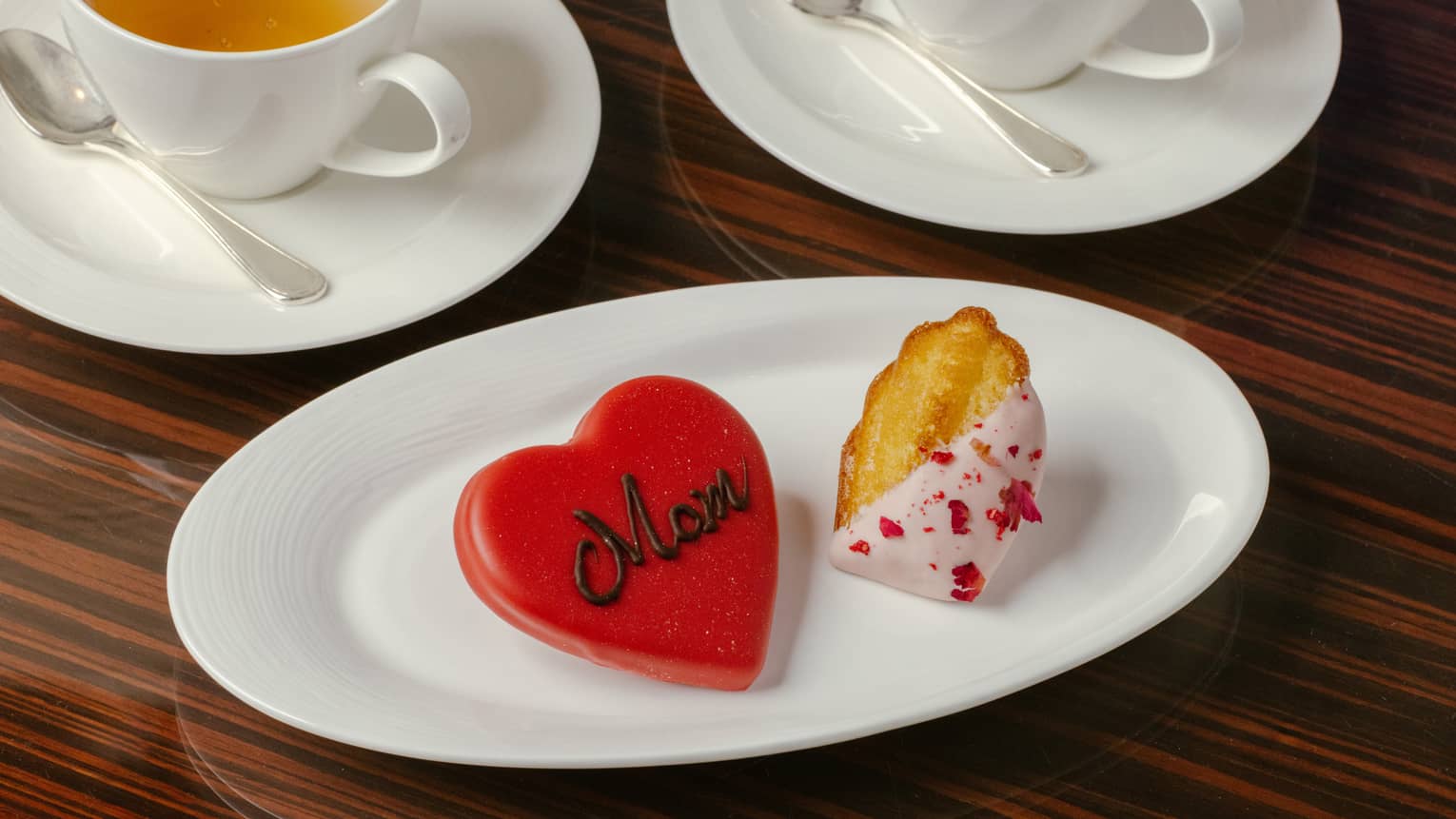 An oval plate holds two cookies. One is red and heart-shaped and has Mom written on it in icing. The other is a golden brown, partially dipped in pinkish-white icing with red sprinkles.