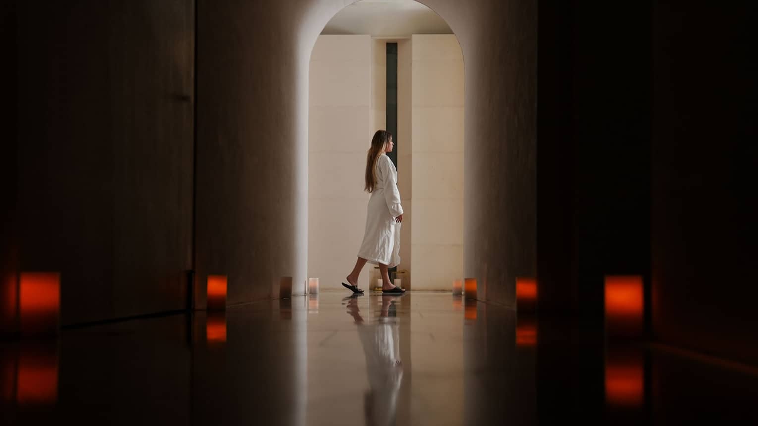 Person with long dark hair wearing a white spa robe walks through a dark hallway lit by eight dim red lanterns