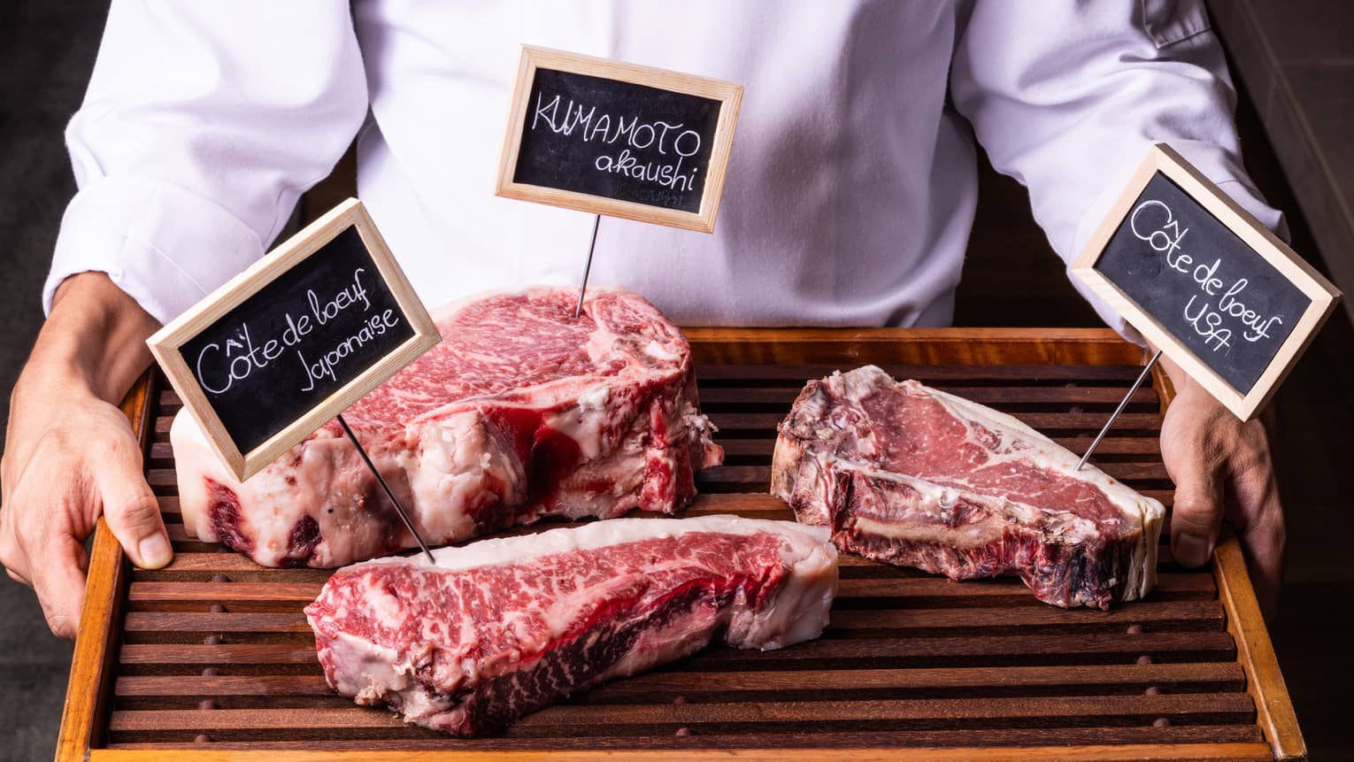 Close-up view of a wood tray with three labelled cuts of well-marbled beef, held by person in white chef's coat.
