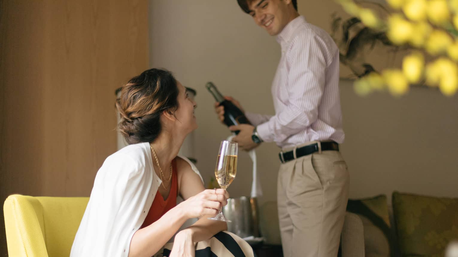 Person smiling and holding a glass of champagne while another person opens a bottle