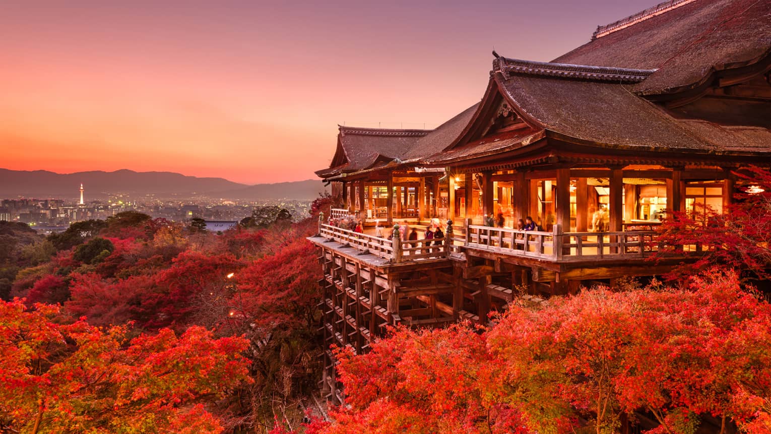 Massive temple on stilts under a pink-hued sky, people on patio admire red-leafed trees, distant city skyline and mountains.