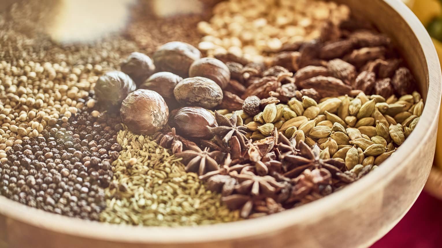 Wooden bowl of Indian spices including whole nutmeg, star anise, cardamom pods and black mustard seeds