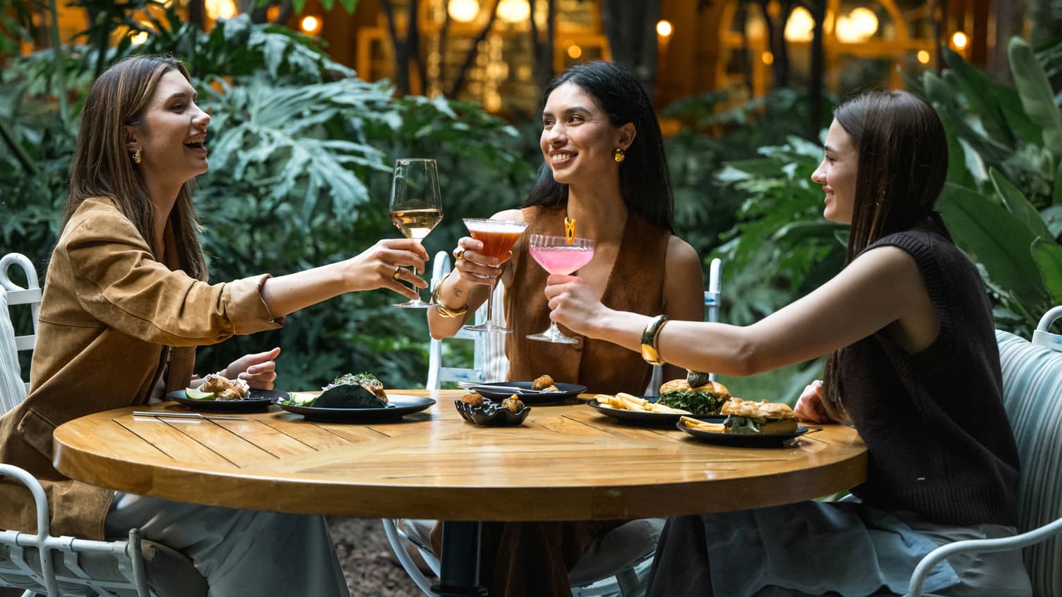 Three guests sit around a round wood table in an outdoor dining space and raise their glasses in a toast.