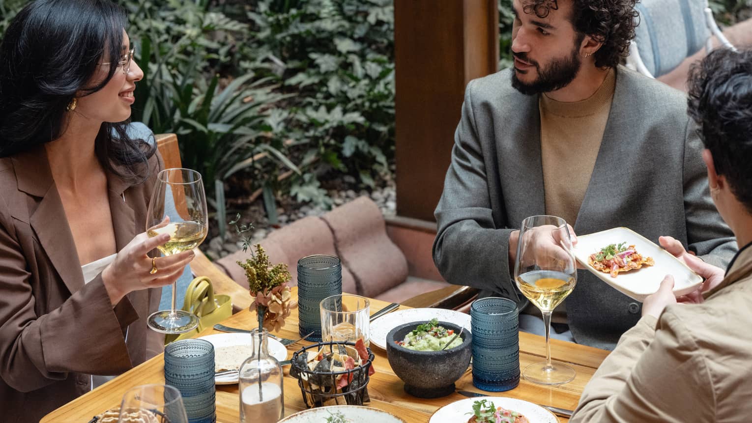 Three people dine at a table set with plates of food and stemware with drinks. Verdant greenery is in the background.