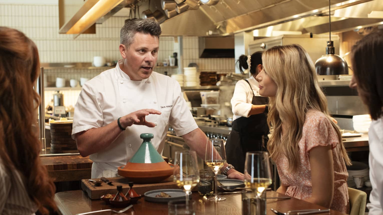 Three guests sitting at a dining table in the kitchen while the chef talks to them.
