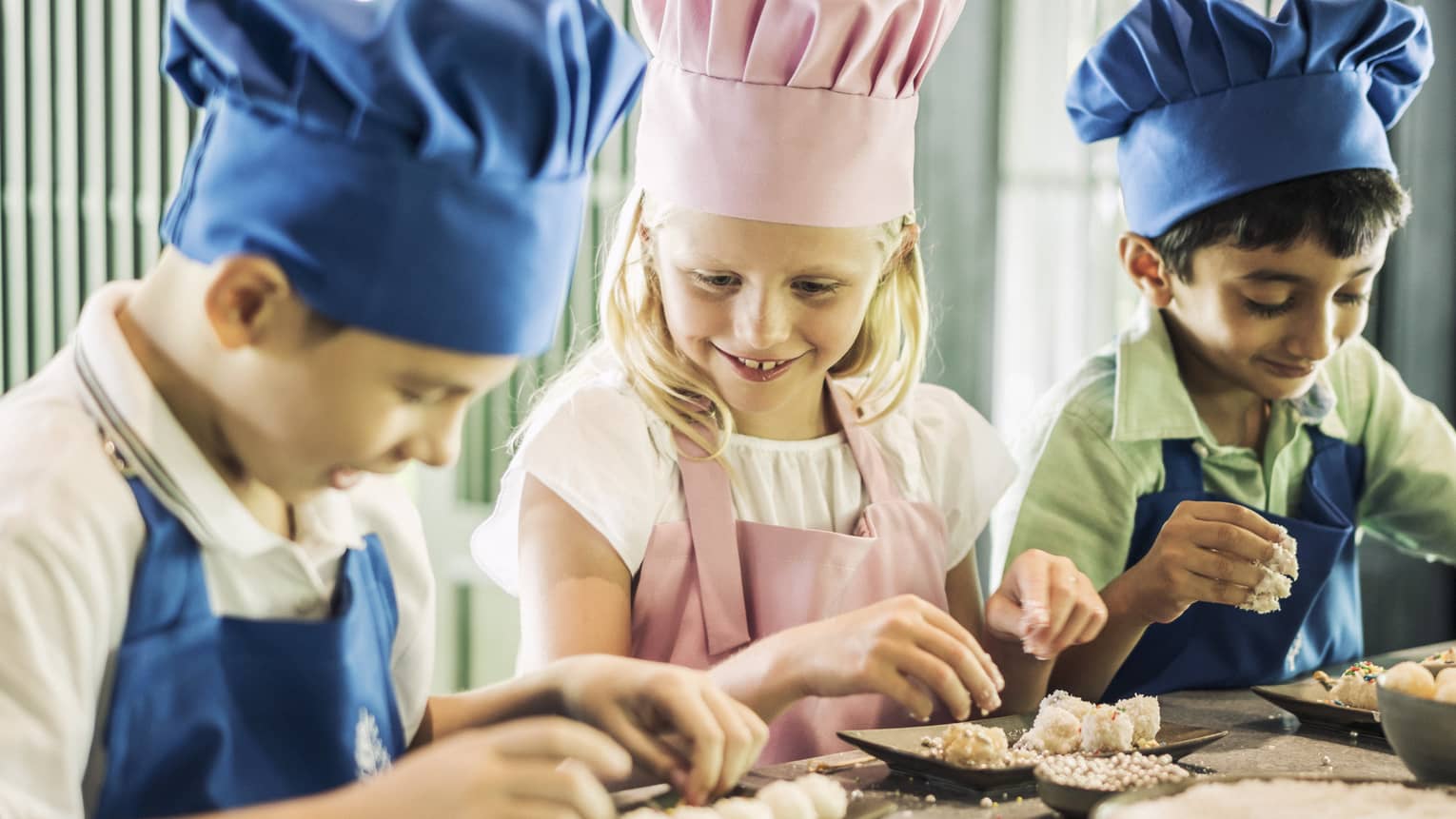 Three children in chef hats, aprons roll pastries at counter 