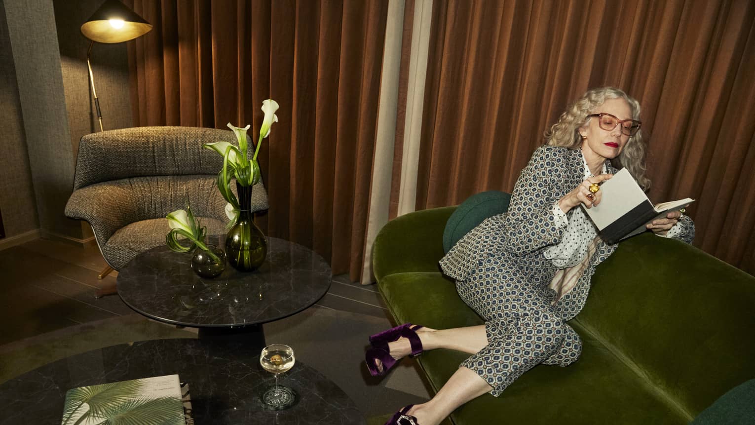 Stylish person with long gray hair wearing a patterned suit and purple heels sitting on a green sofa while reading a book
