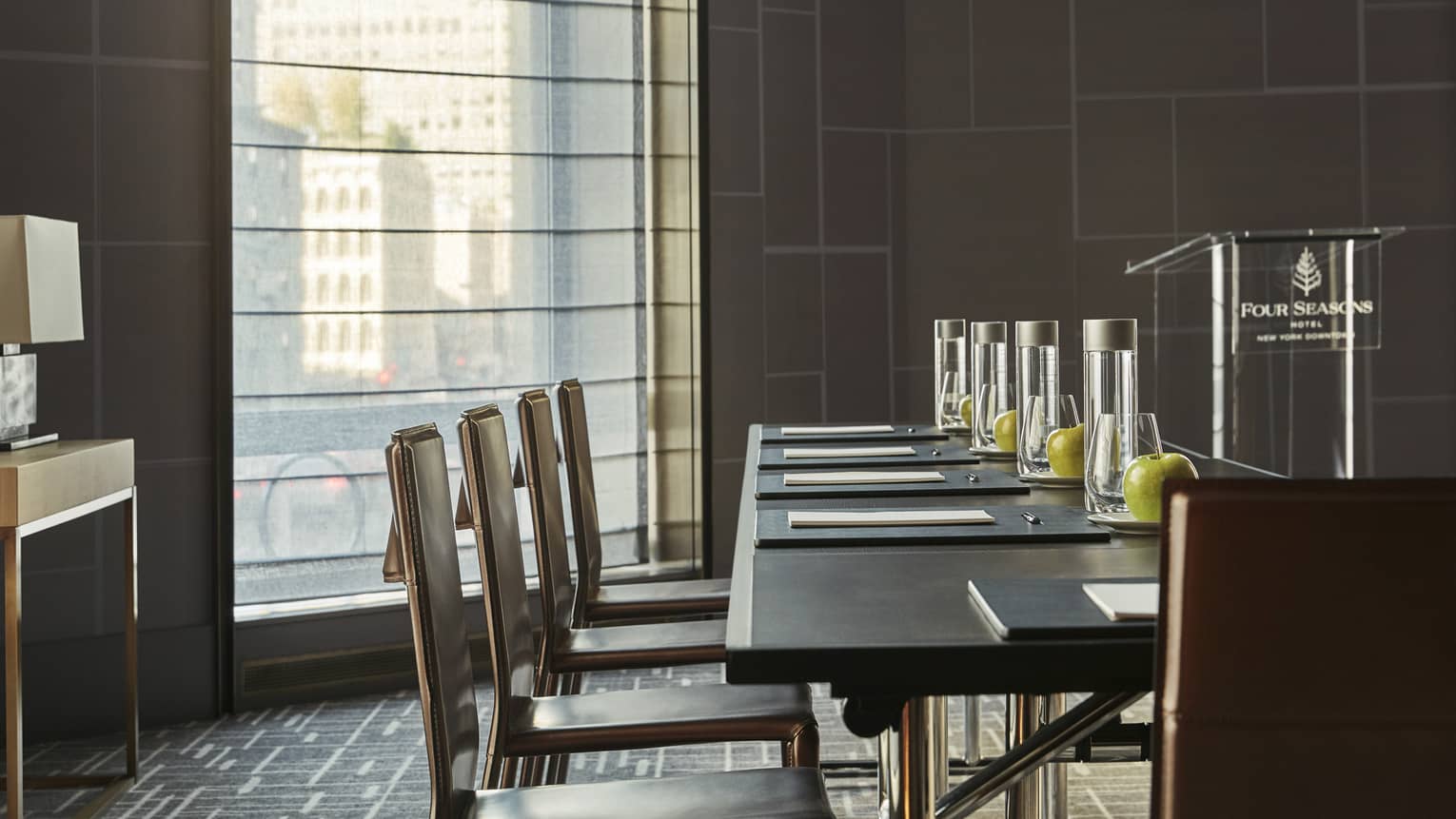 Dark table with chairs, stationery, bottles of water, green apples in light-filled meeting room with podium