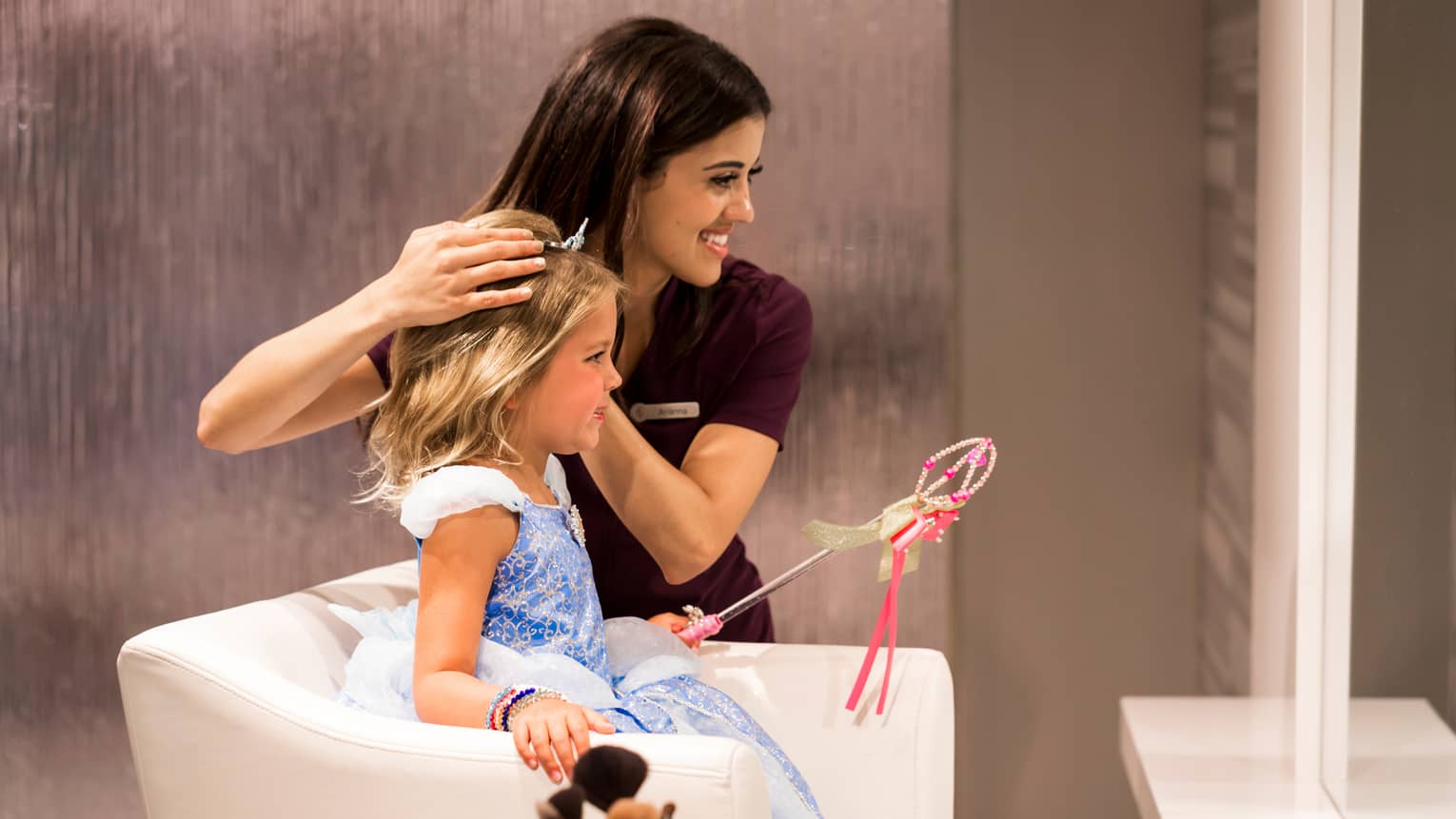 Little girl dressed as princess sits in chair, hotel staff places tiara in her hair