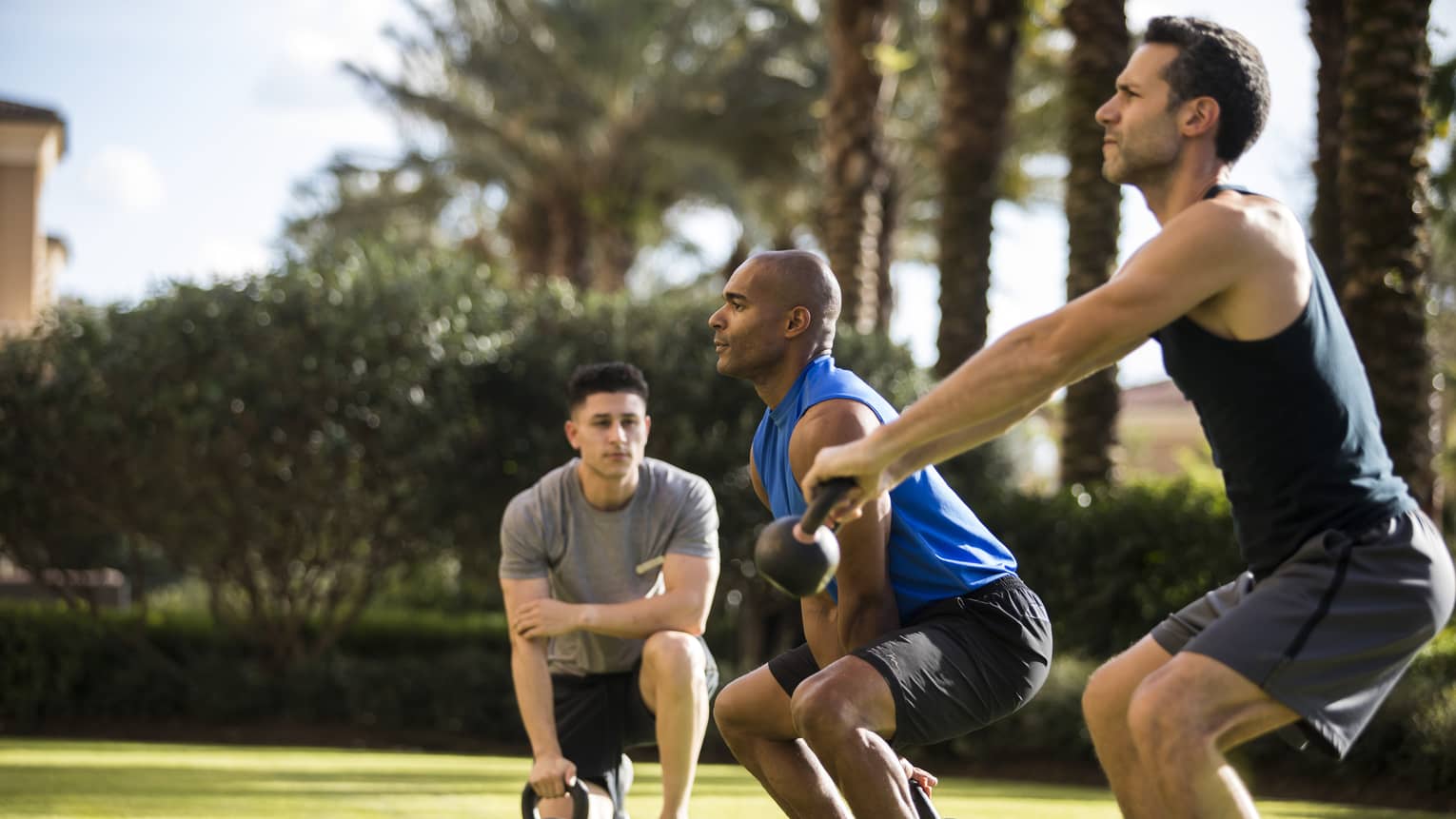 Four Seasons trainer watches as two men squat, lift kettlebell weights on lawn