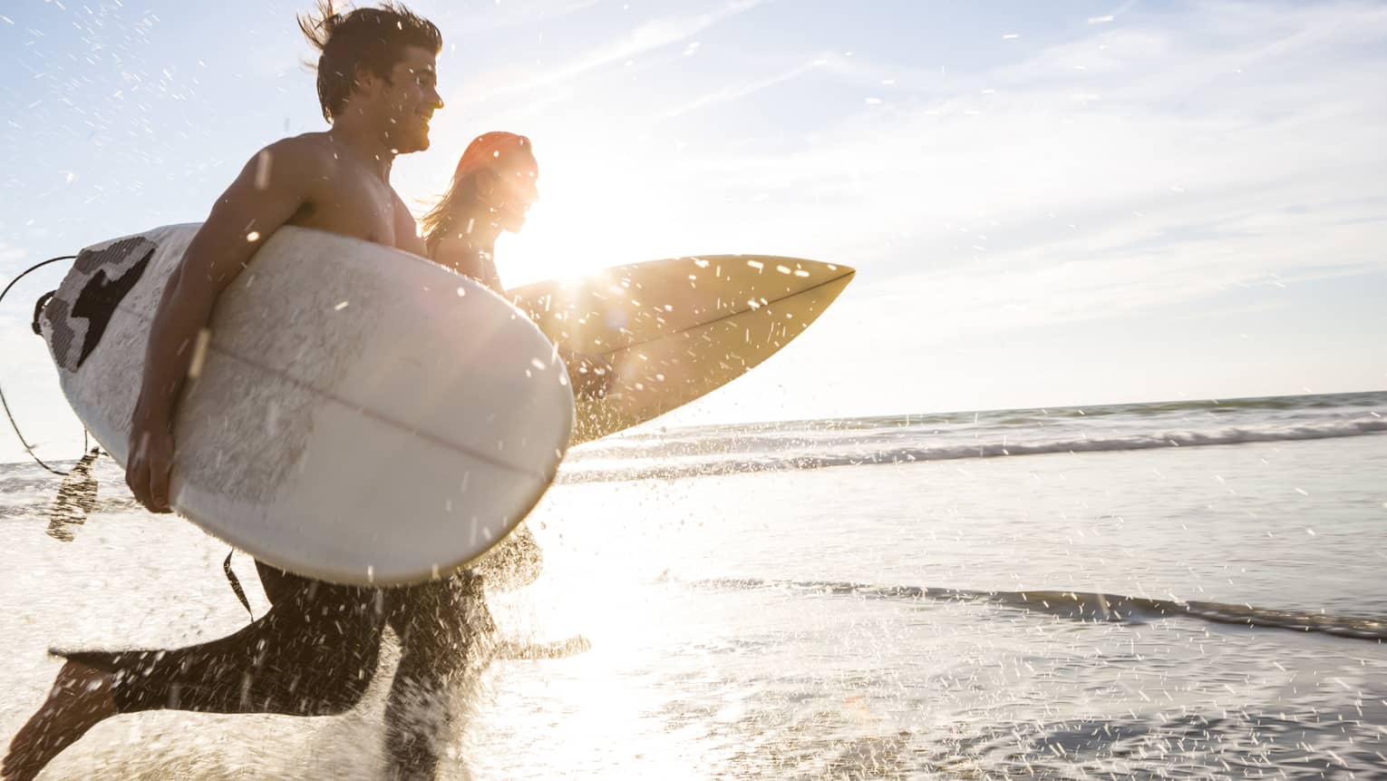 Couple hold surfboards, run across beach tide at sunrise