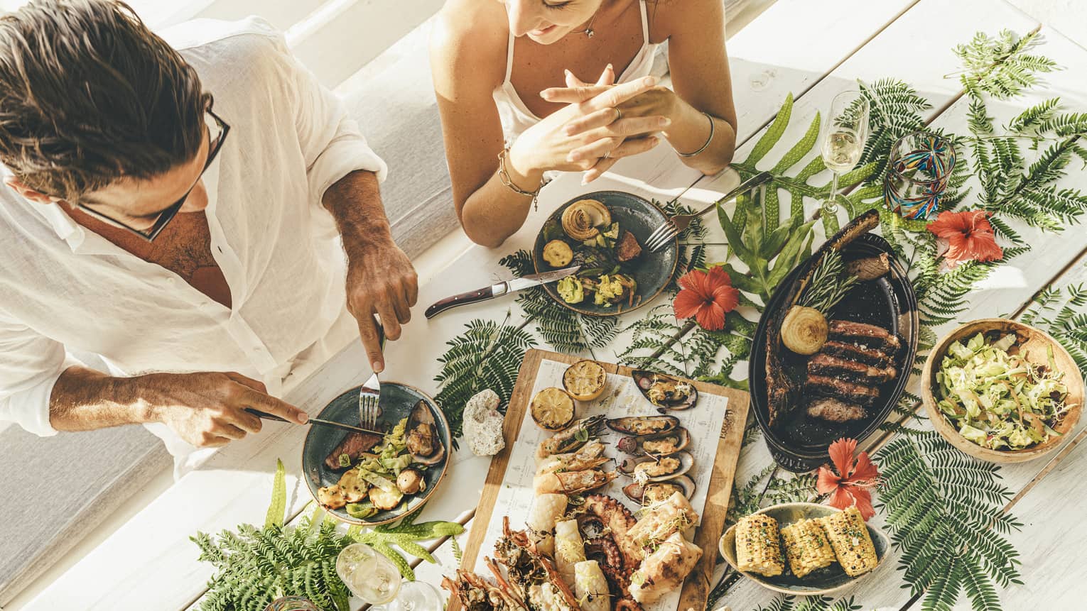 A couple at a table of seafood canapés, grilled meat, corn and champagne flutes, adorned with palm fronds and red hibiscus.