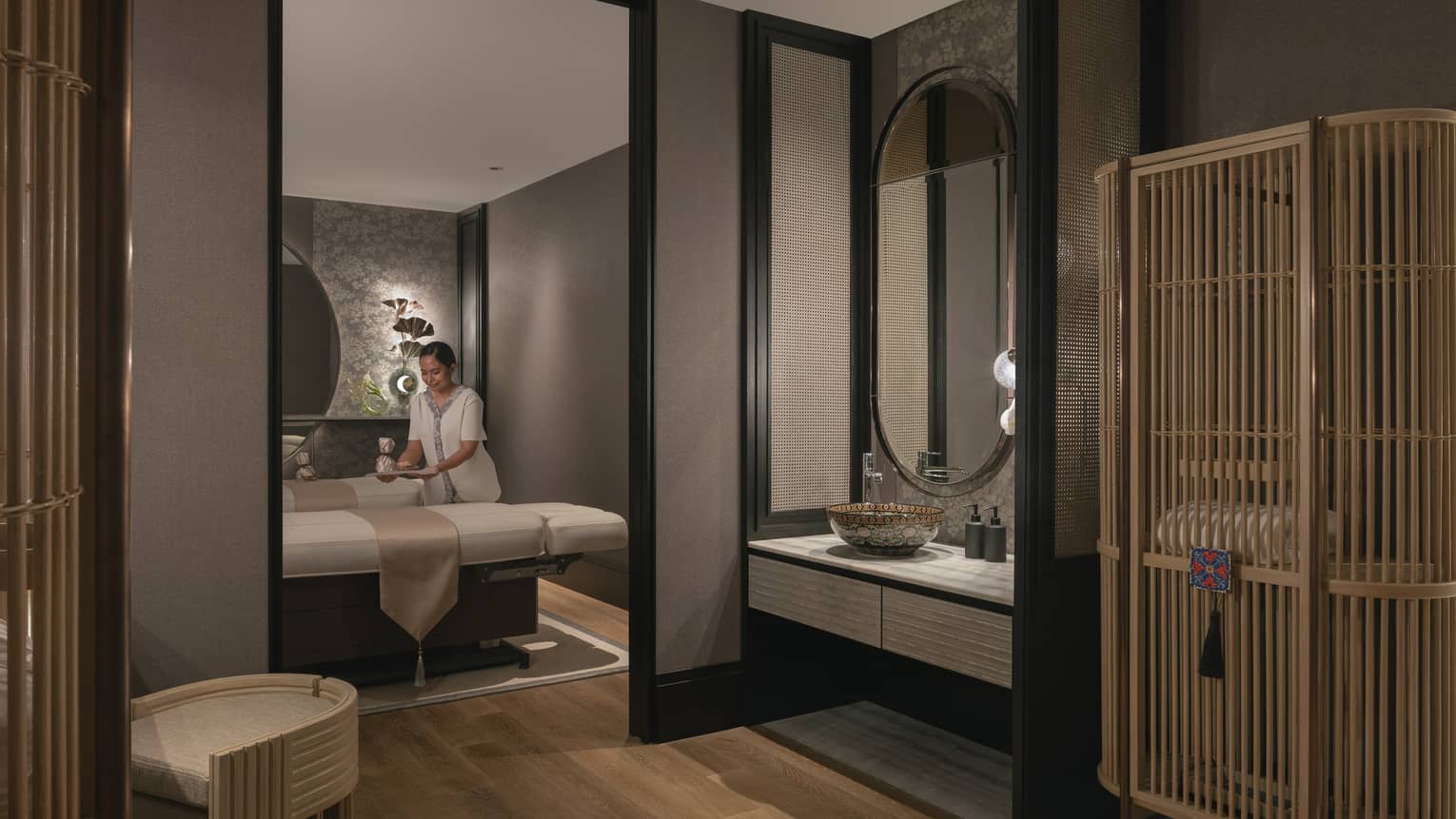 A tray is gently placed on one of two massage tables in a warmly lit spa room with soft grey walls and natural wood décor