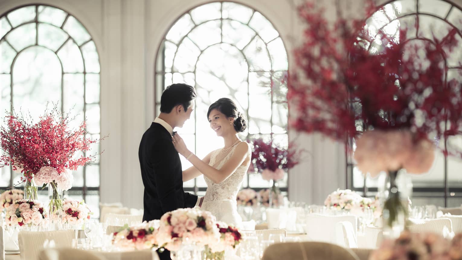 Woman in wedding dress adjusts groom's bow tie in light filled banquet room with pink flowers