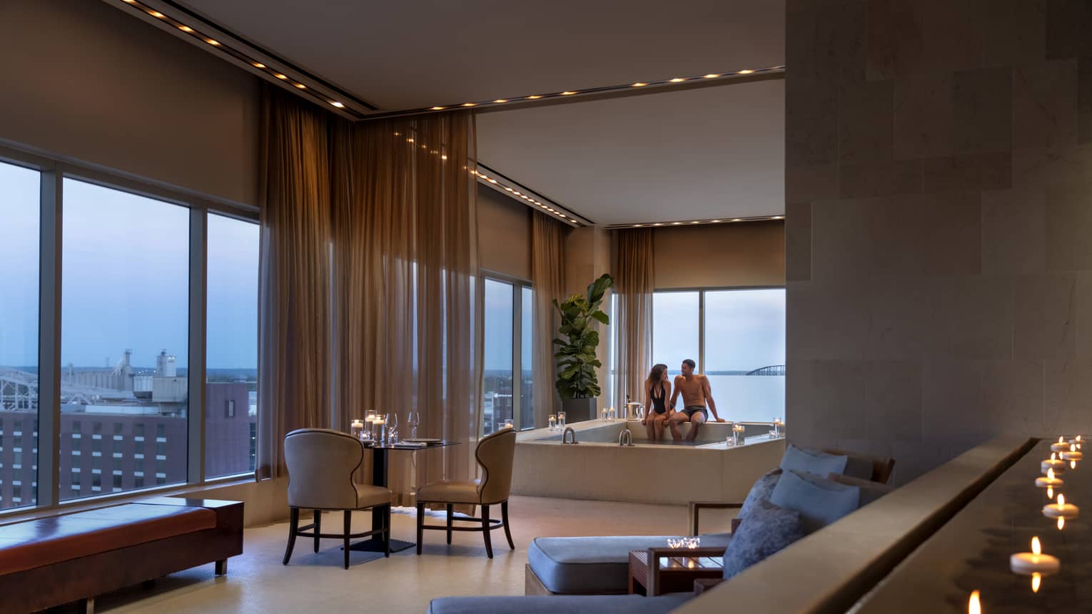 A couple enjoying a candlelit hot tub in a luxury hotel spa, with elegant seating, soft lighting and large windows offering a view of a distant bridge
