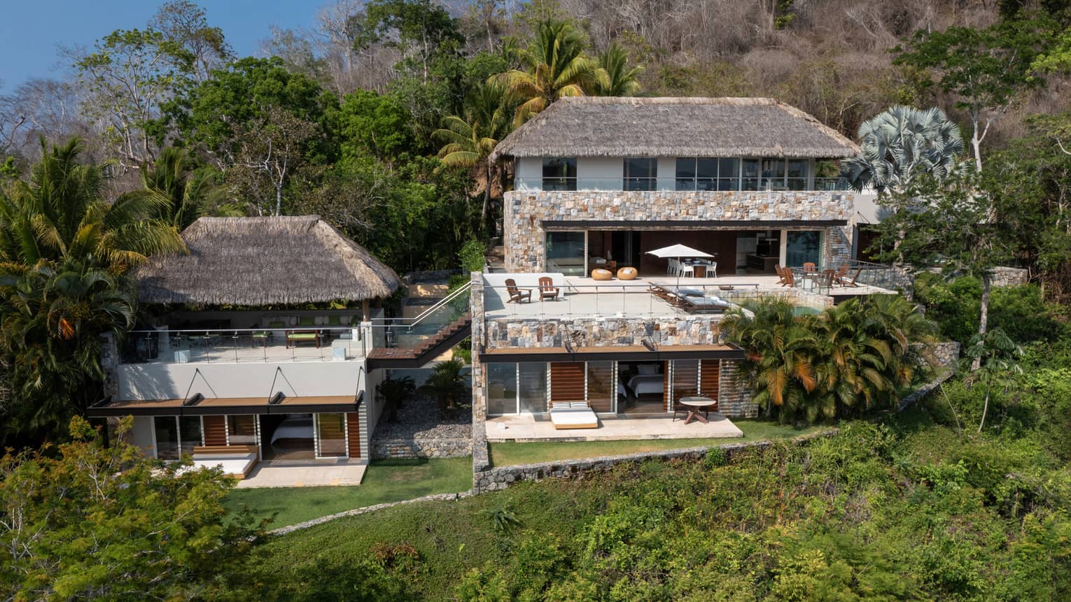 A multi-level luxury villa nestled on a hillside, featuring thatched roofs, stone walls, spacious terraces and lush tropical greenery surrounding the property
