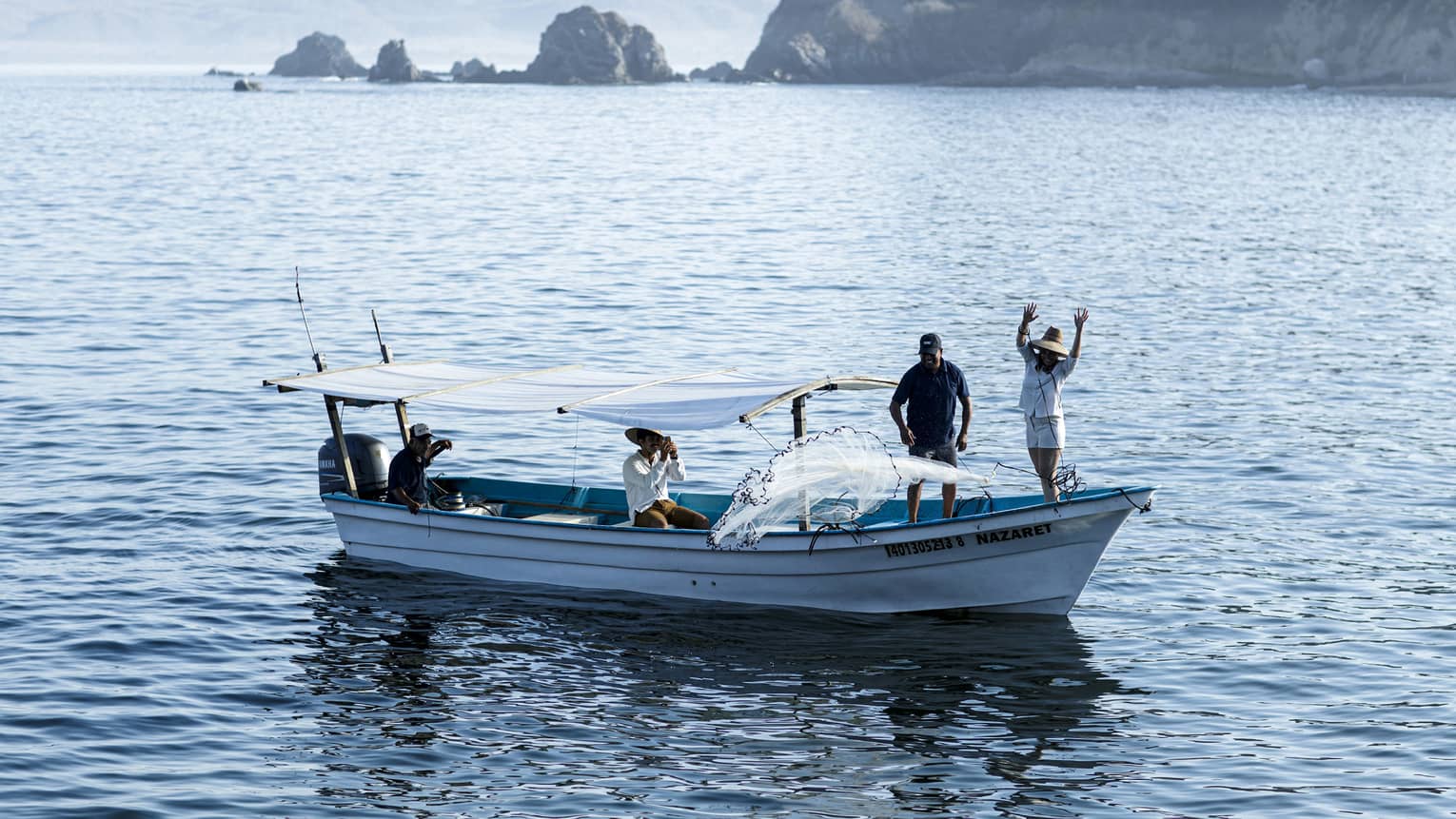 Side view of a boat with two people at the bow casting a net into the water, two others seated under a canopy near the stern.