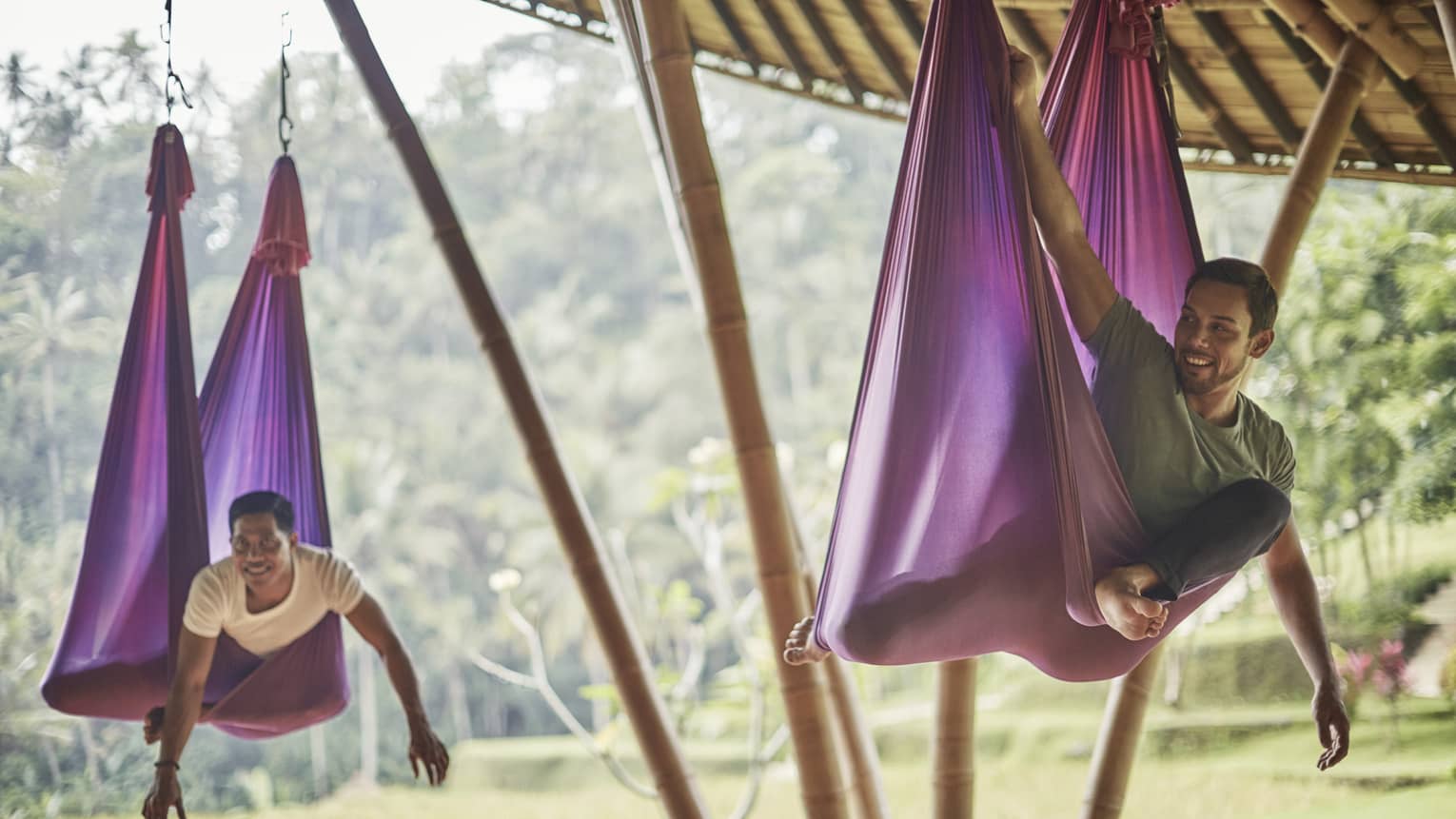Two men hang from large purple yoga hammocks under wood pavillion