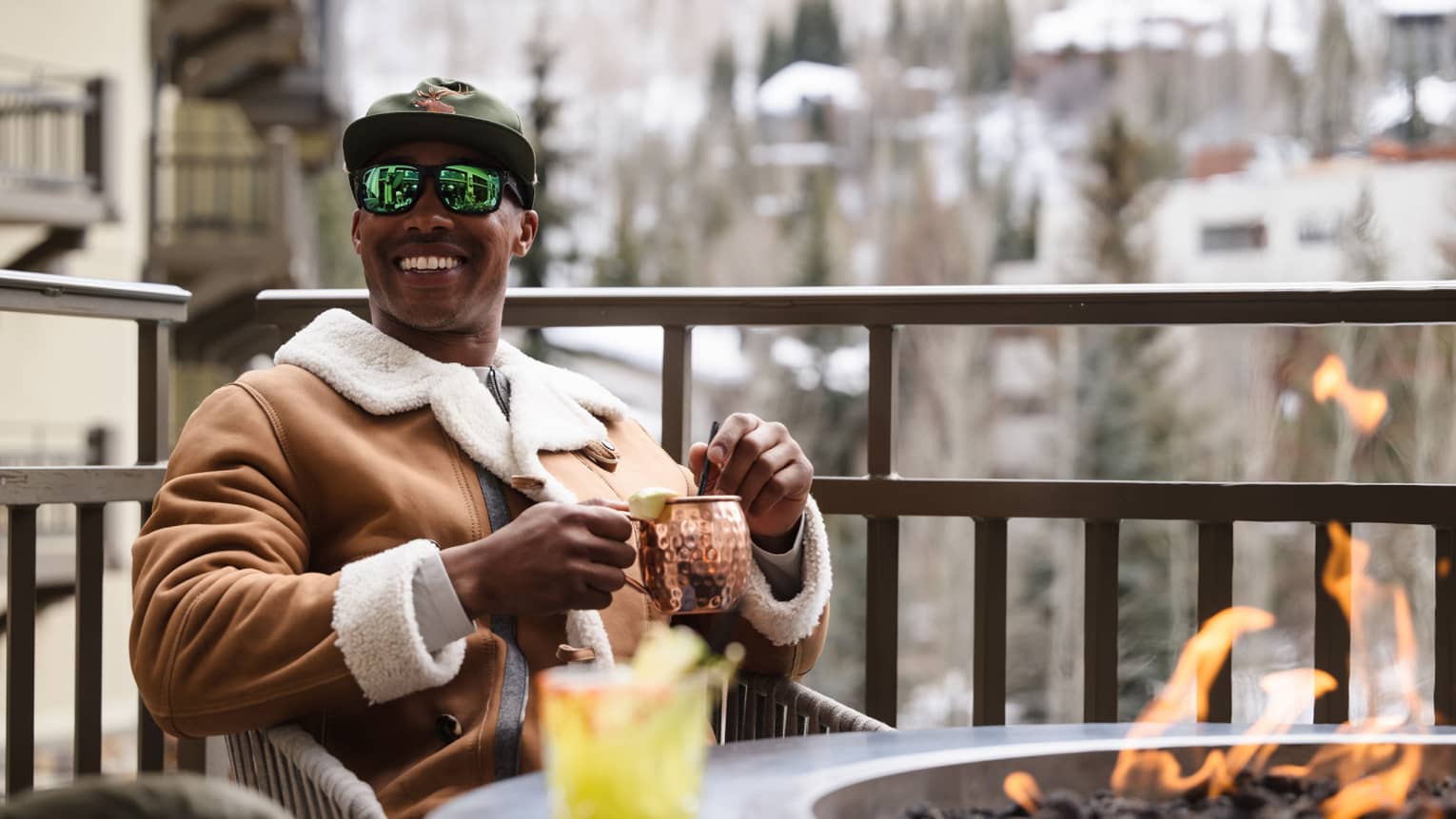 Guest wearing a jacket and holding a mug while sitting near a fire pit.