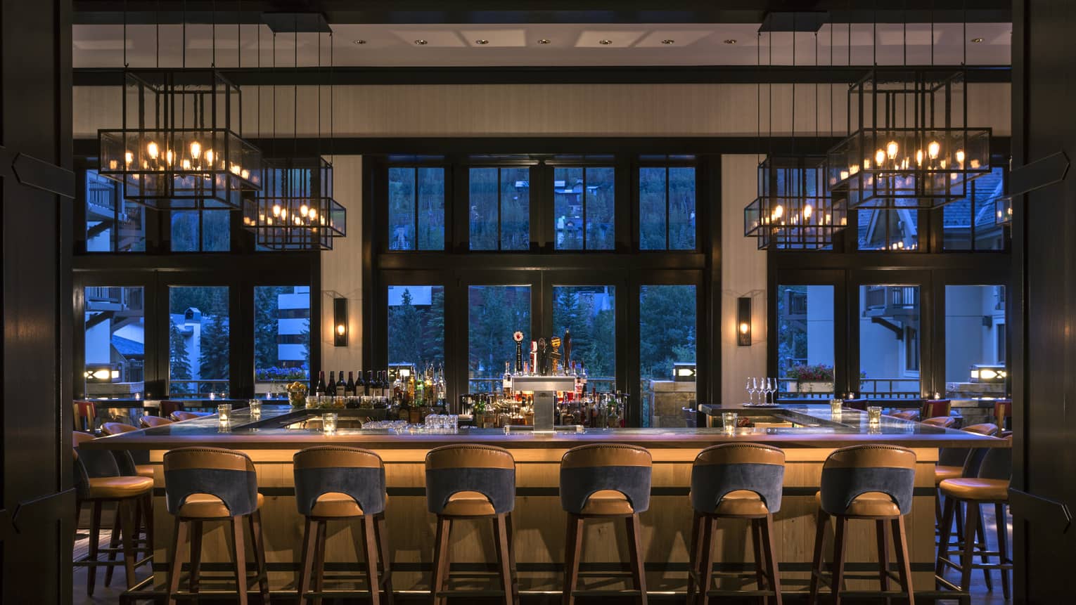 Dimly-lit bar at dusk lined with retro-style wood stools, lantern chandeliers above, large windows