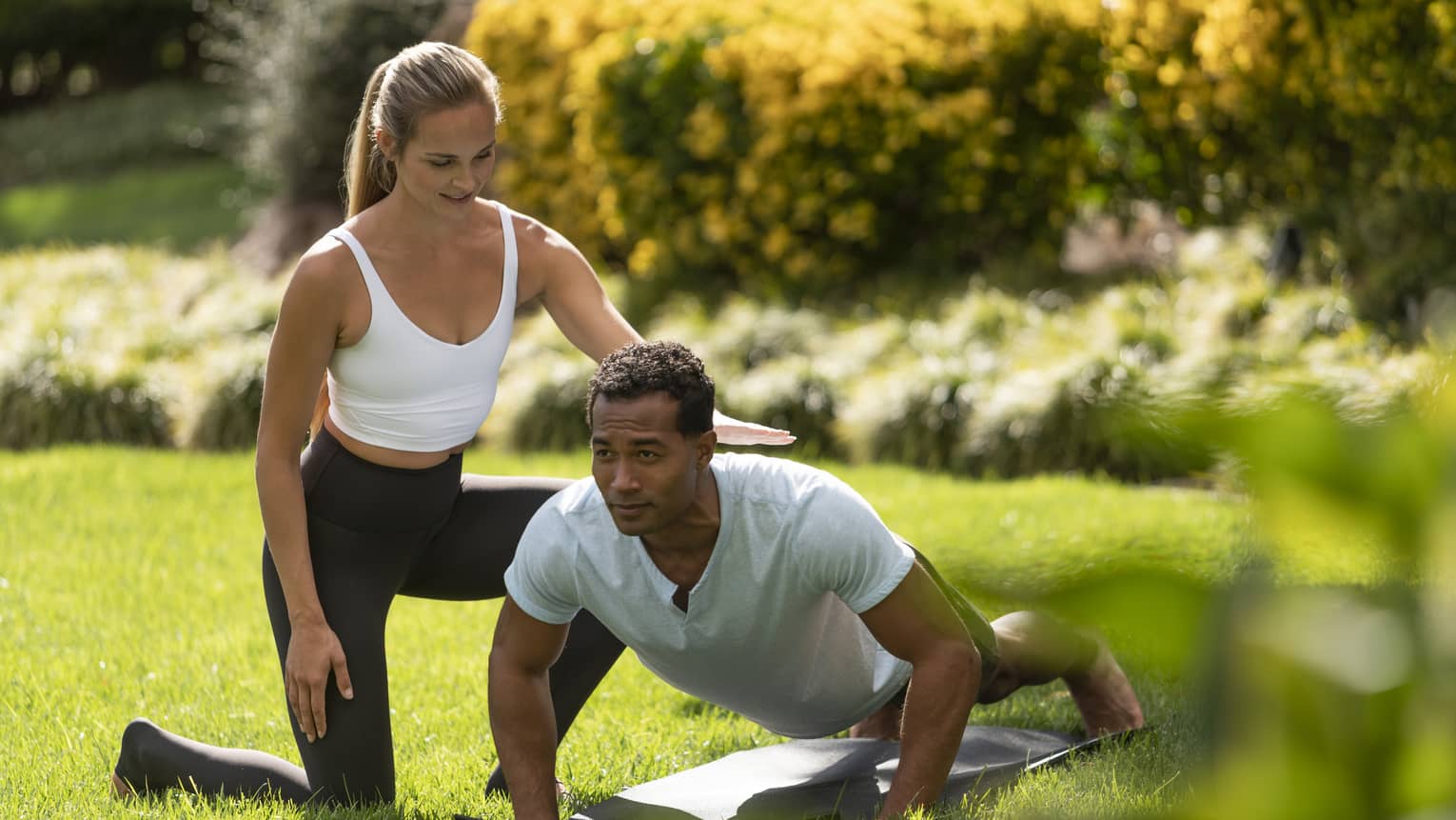 A yoga instructor helping a man get into a yoga position outside on very green grass.