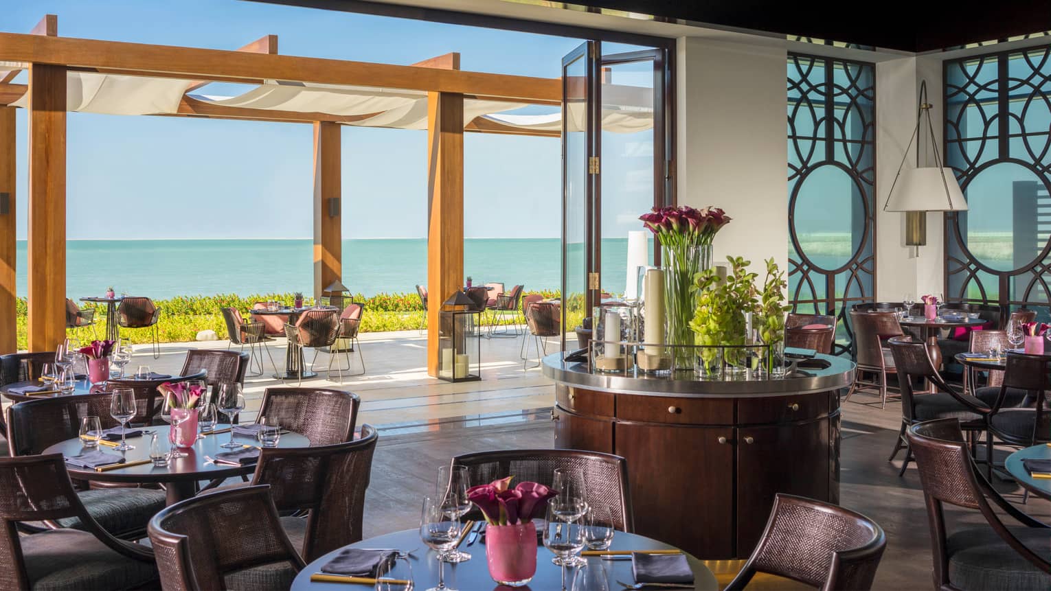 Sea Fu indoor outdoor dining room with round tables and glass wall open to patio with awnings