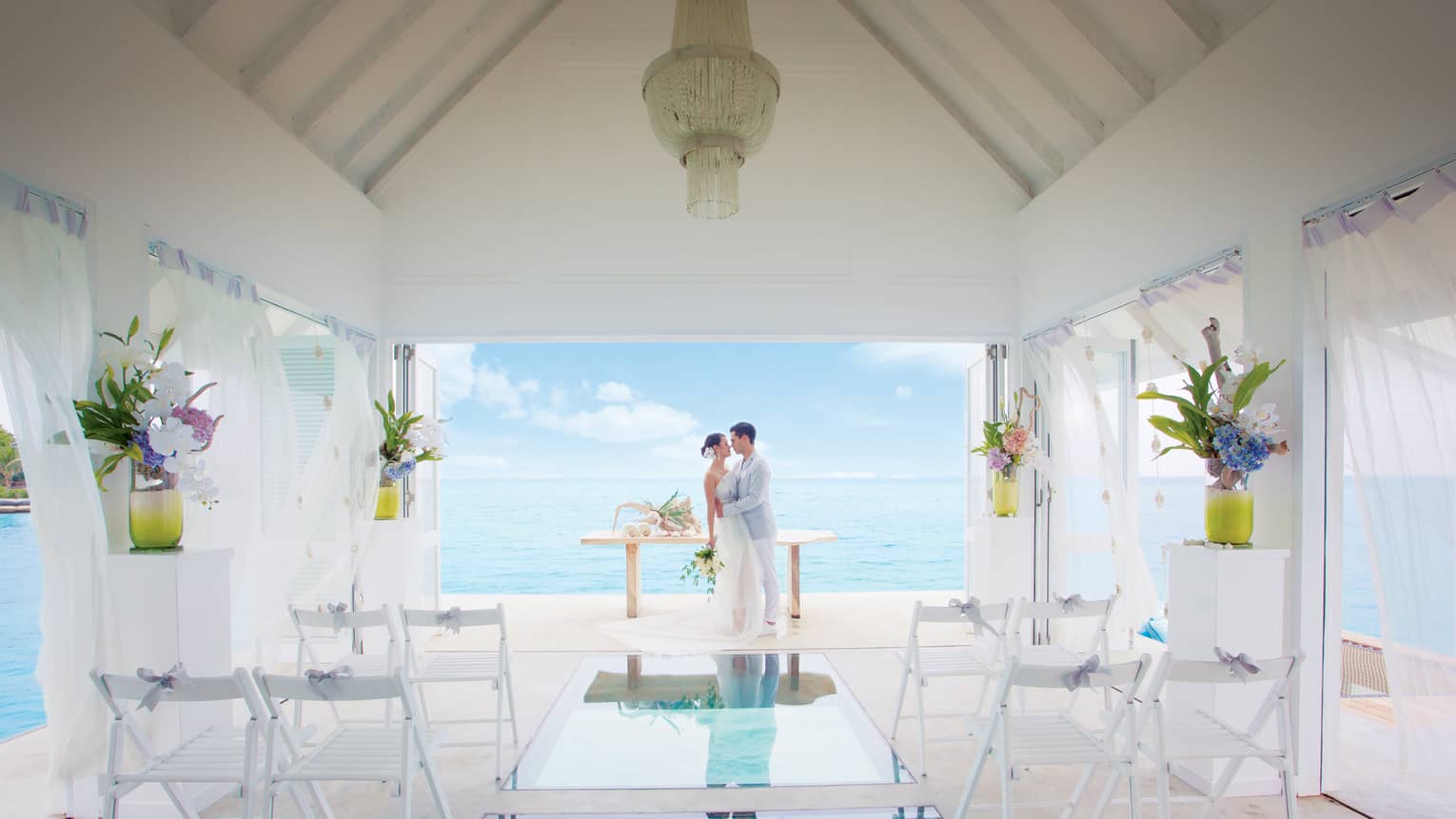Bride and groom embrace at edge of small white bungalow with glass floors looking down into blue lagoon