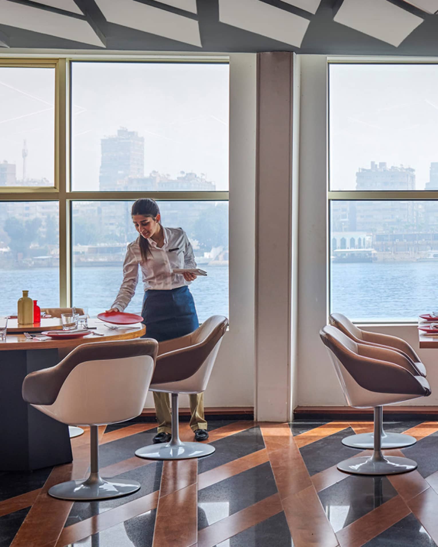 Contemporary restaurant with server on left, rectangular tables, lattice floor, Nile River views
